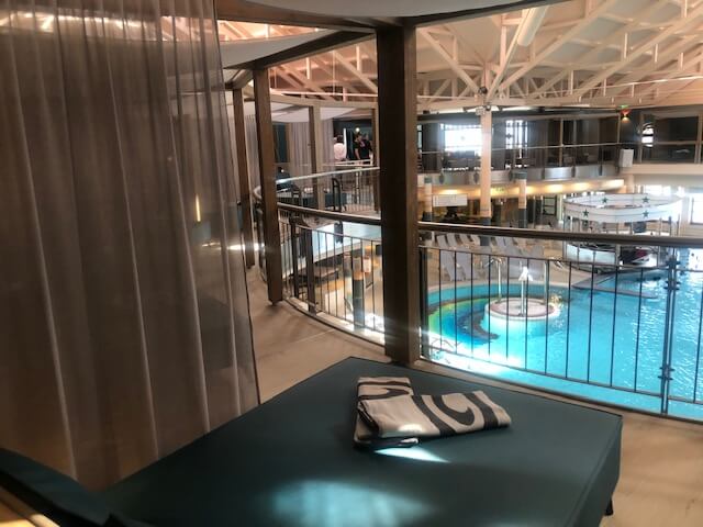 Ein großer Innenpoolbereich in einem Spa mit hellblauem Wasser und mehreren runden Becken. In der Mitte des Hauptbeckens befindet sich eine runde Poolbar mit einer weißen Überdachung und grünen Sternen. Der Bereich ist von einem zweistöckigen Balkon mit schwarzen Geländern umgeben. Im Hintergrund sind zahlreiche Liegestühle, Sitzgelegenheiten und ein Essbereich zu sehen. Die Decke ist hoch und mit weißen Stahlträgern versehen, die den Raum offen und geräumig wirken lassen. Große Fenster auf der rechten Seite lassen viel Tageslicht in den Raum.