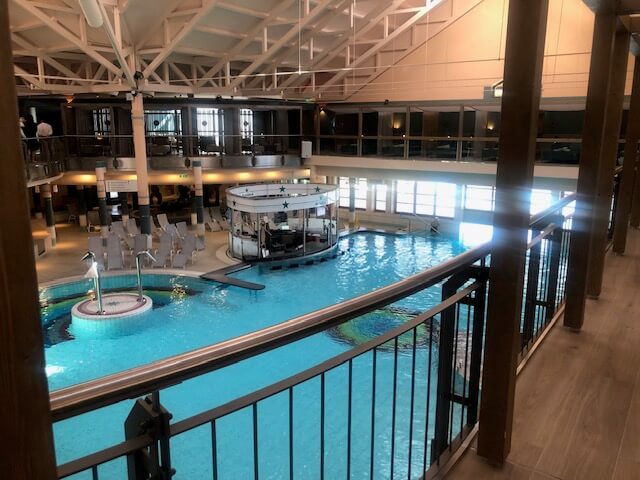 Ein großer Innenpoolbereich in einem Spa mit hellblauem Wasser und mehreren runden Becken. In der Mitte des Hauptbeckens befindet sich eine runde Poolbar mit einer weißen Überdachung und grünen Sternen. Der Bereich ist von einem zweistöckigen Balkon mit schwarzen Geländern umgeben. Im Hintergrund sind zahlreiche Liegestühle, Sitzgelegenheiten und ein Essbereich zu sehen. Die Decke ist hoch und mit weißen Stahlträgern versehen, die den Raum offen und geräumig wirken lassen. Große Fenster auf der rechten Seite lassen viel Tageslicht in den Raum.