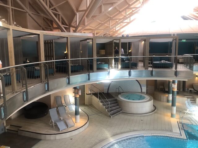 Ein großer Innenpoolbereich in einem Spa mit hellblauem Wasser und mehreren runden Becken. In der Mitte des Hauptbeckens befindet sich eine runde Poolbar mit einer weißen Überdachung und grünen Sternen. Der Bereich ist von einem zweistöckigen Balkon mit schwarzen Geländern umgeben. Im Hintergrund sind zahlreiche Liegestühle, Sitzgelegenheiten und ein Essbereich zu sehen. Die Decke ist hoch und mit weißen Stahlträgern versehen, die den Raum offen und geräumig wirken lassen. Große Fenster auf der rechten Seite lassen viel Tageslicht in den Raum.