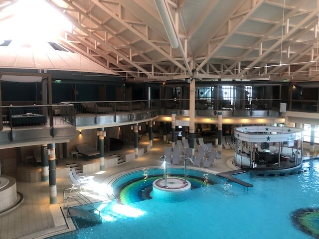 Ein großer Innenpoolbereich in einem Spa mit hellblauem Wasser und mehreren runden Becken. In der Mitte des Hauptbeckens befindet sich eine runde Poolbar mit einer weißen Überdachung und grünen Sternen. Der Bereich ist von einem zweistöckigen Balkon mit schwarzen Geländern umgeben. Im Hintergrund sind zahlreiche Liegestühle, Sitzgelegenheiten und ein Essbereich zu sehen. Die Decke ist hoch und mit weißen Stahlträgern versehen, die den Raum offen und geräumig wirken lassen. Große Fenster auf der rechten Seite lassen viel Tageslicht in den Raum.