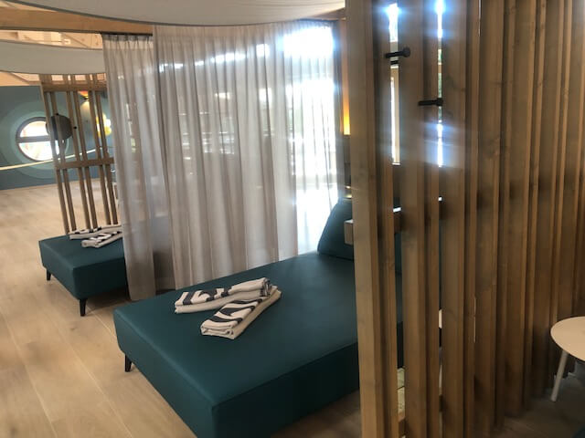 Ein stilvoller Ruhebereich in einem Spa mit türkisfarbenen Liegen, die durch vertikale Holzpaneele voneinander getrennt sind. Auf den Liegen liegen sorgfältig gefaltete Handtücher. Die Paneele bieten eine teilweise Abgrenzung, während durchsichtige Vorhänge zusätzliche Privatsphäre ermöglichen. Der Boden besteht aus hellen Holzdielen, die den Raum freundlich und einladend wirken lassen. Im Hintergrund sind runde Fenster zu sehen, die natürliches Licht in den Raum lassen und für eine entspannte Atmosphäre sorgen.