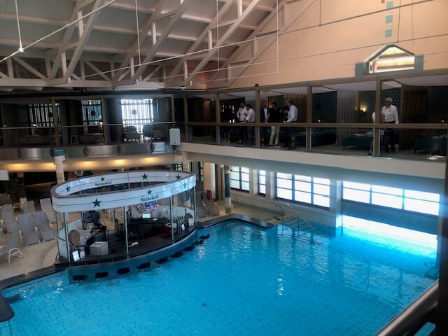 Ein großer Innenpoolbereich in einem Spa mit hellblauem Wasser und mehreren runden Becken. In der Mitte des Hauptbeckens befindet sich eine runde Poolbar mit einer weißen Überdachung und grünen Sternen. Der Bereich ist von einem zweistöckigen Balkon mit schwarzen Geländern umgeben. Im Hintergrund sind zahlreiche Liegestühle, Sitzgelegenheiten und ein Essbereich zu sehen. Die Decke ist hoch und mit weißen Stahlträgern versehen, die den Raum offen und geräumig wirken lassen. Große Fenster auf der rechten Seite lassen viel Tageslicht in den Raum.