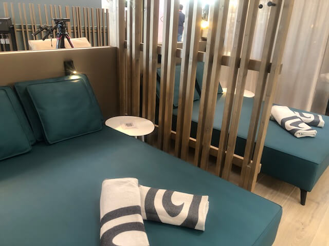 Ein stilvoller Ruhebereich in einem Spa mit türkisfarbenen Liegen, die durch vertikale Holzpaneele voneinander getrennt sind. Auf den Liegen liegen sorgfältig gefaltete Handtücher. Die Paneele bieten eine teilweise Abgrenzung, während durchsichtige Vorhänge zusätzliche Privatsphäre ermöglichen. Der Boden besteht aus hellen Holzdielen, die den Raum freundlich und einladend wirken lassen. Im Hintergrund sind runde Fenster zu sehen, die natürliches Licht in den Raum lassen und für eine entspannte Atmosphäre sorgen.