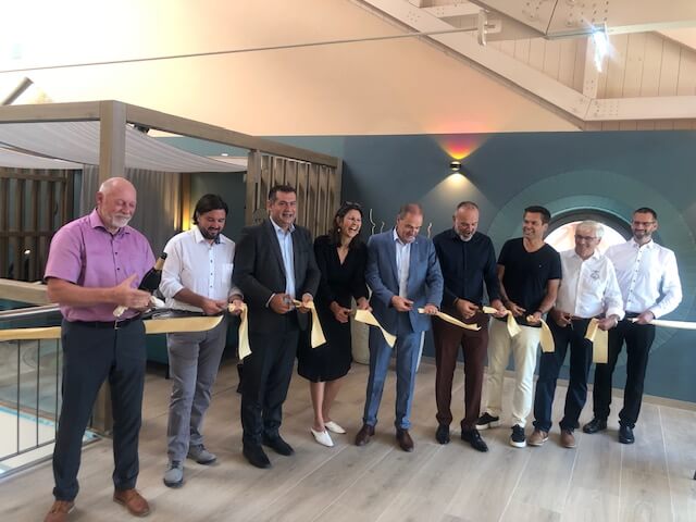 Offizielle Eröffnung des AVITA NEXT LEVEL: Geschäftsführer Peter Prisching (4.v.r.) und acht weitere Personen stehen nebeneinander und halten gemeinsam ein breites Band für die Zeremonie. Die Gruppe ist unterschiedlich gekleidet, von formellen Anzügen bis zu Freizeitkleidung. Der Raum im Hintergrund ist modern gestaltet, mit türkisfarbenen Wänden, runden Fenstern und hellen Holzböden. Vertikale Holzpaneele und durchsichtige Vorhänge trennen verschiedene Bereiche, während gedämpfte Beleuchtung für eine angenehme Atmosphäre sorgt.