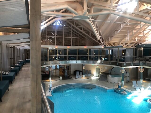 Ein großer Innenpoolbereich in einem Spa mit hellblauem Wasser und mehreren runden Becken. In der Mitte des Hauptbeckens befindet sich eine runde Poolbar mit einer weißen Überdachung und grünen Sternen. Der Bereich ist von einem zweistöckigen Balkon mit schwarzen Geländern umgeben. Im Hintergrund sind zahlreiche Liegestühle, Sitzgelegenheiten und ein Essbereich zu sehen. Die Decke ist hoch und mit weißen Stahlträgern versehen, die den Raum offen und geräumig wirken lassen. Große Fenster auf der rechten Seite lassen viel Tageslicht in den Raum.