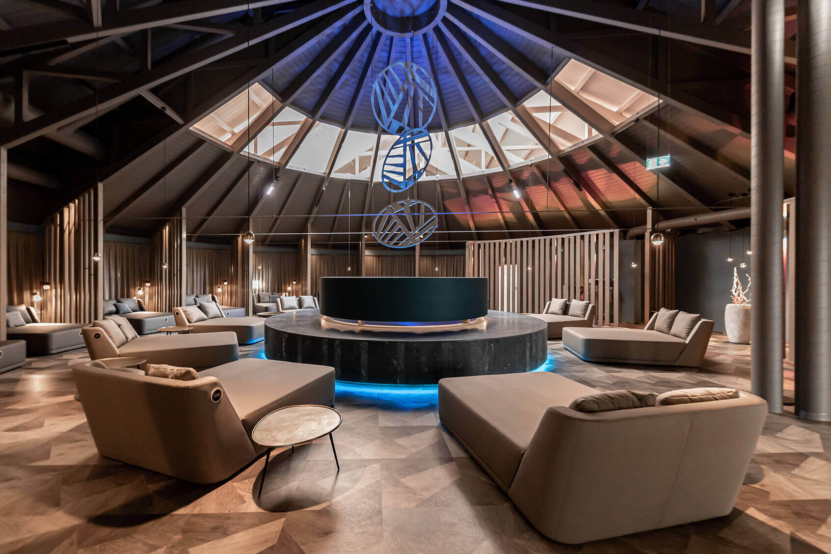 Ein eleganter Ruhebereich mit hoher, gewölbter Decke und modernen Designelementen. In der Mitte des Raumes befindet sich ein großer, runder Tisch mit blauer Beleuchtung am Sockel. Darum herum sind beige Liegen in einem Kreis angeordnet, jede mit Kissen und kleinen Beistelltischen. Die Decke ist aus dunklem Holz und wird von stilvollen Hängeleuchten beleuchtet. Große, leuchtende Kunstinstallationen hängen von der Decke herab. Im Hintergrund sind Holzlamellenwände und Vorhänge zu sehen, die eine gemütliche und ruhige Atmosphäre schaffen. ©Avita Resort