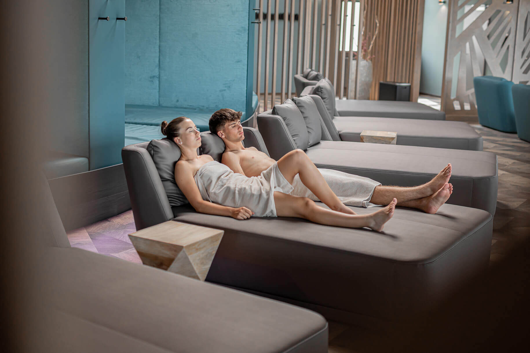 Ein Mann und eine Frau liegen entspannt auf einer grauen Liege in einem modernen Spa-Bereich. Beide sind in weiße Handtücher gewickelt. Die Frau hat die Augen geschlossen und lehnt sich an den Mann, der ebenfalls entspannt daliegt. Im Hintergrund sind weitere graue Liegen zu sehen, die in Reihen angeordnet sind. ©Avita Resort