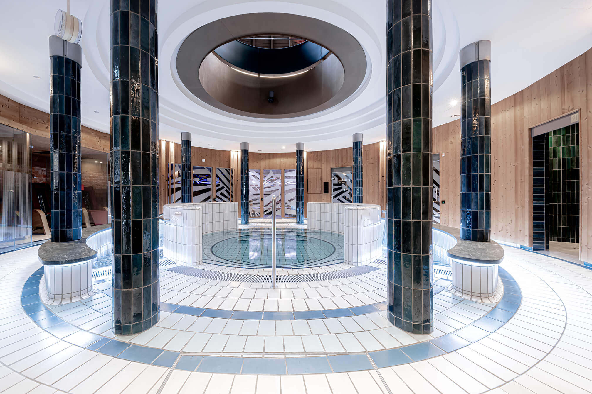 Ein eleganter Spa-Bereich mit einem runden Pool in der Mitte, umgeben von vier dunkelblau gefliesten Säulen. Der Boden und die Wände des Pools sind mit weißen Fliesen bedeckt, und es führen Stufen in den Pool hinunter. Der Raum hat eine hohe Decke mit einem runden Oberlicht in der Mitte, das Tageslicht hereinlässt. Die Wände sind aus Holz und teilweise mit modernen Kunstinstallationen verziert. Um den Pool herum sind Sitzgelegenheiten mit beleuchteten Sockeln integriert. Der Raum strahlt eine ruhige und luxuriöse Atmosphäre aus. © Avita Resort