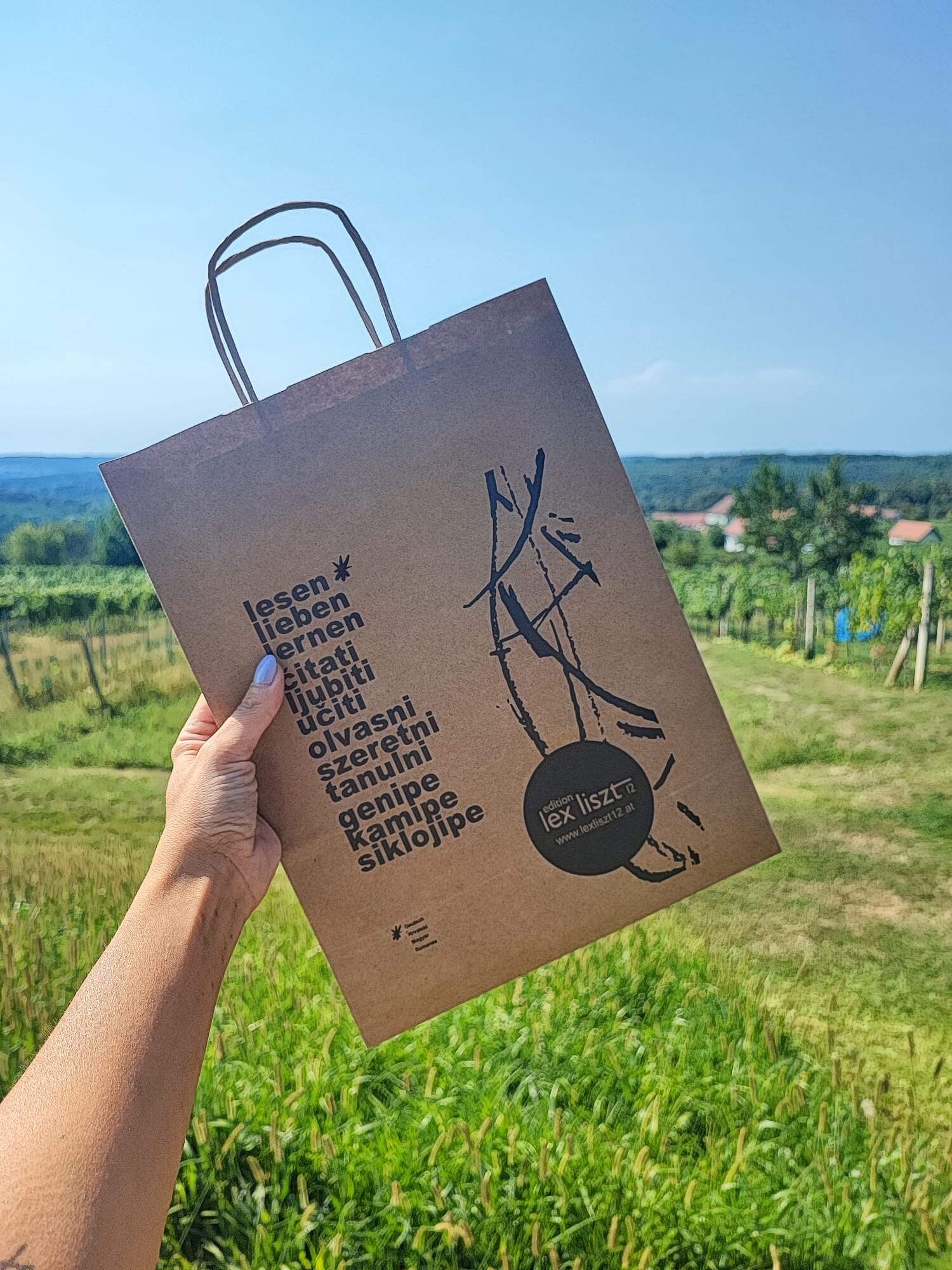 Das Bild zeigt eine Hand, die eine braune Papiertüte in die Höhe hält. Auf der Tüte ist der Schriftzug *"lesen, lieben, lernen"* sowie dessen Übersetzung in verschiedenen Sprachen zu sehen. Darunter befindet sich das Logo der *edition lex liszt 12*. Im Hintergrund erstreckt sich eine grüne Landschaft mit Weinstöcken und einem weiten Blick über Felder und Hügel. Der Himmel ist klar und blau, und die Szene vermittelt eine sommerliche, entspannte Atmosphäre in einer ländlichen Umgebung.