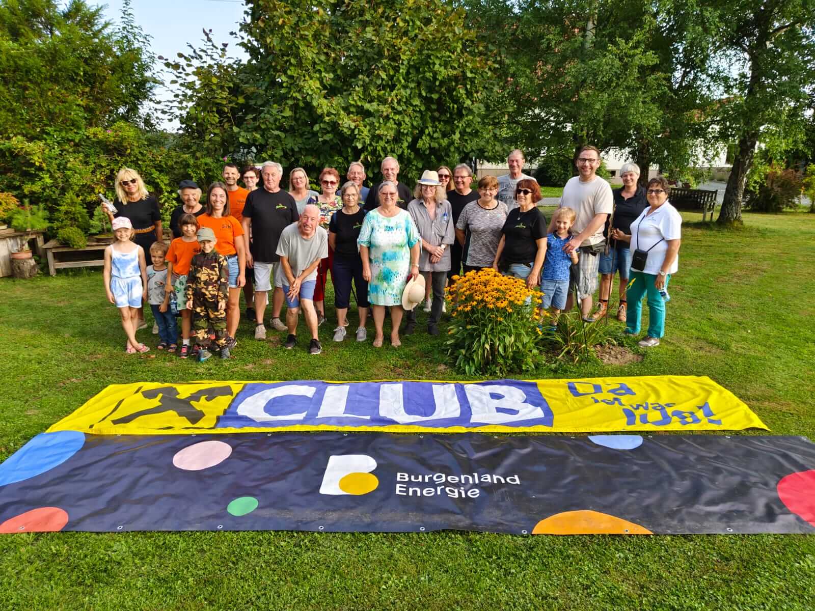 Eine Gruppe von etwa zwanzig Personen steht auf einer Rasenfläche vor einer dichten grünen Hecke. Im Vordergrund sind zwei große Banner auf dem Gras ausgebreitet. Das linke Banner ist gelb und blau mit der Aufschrift "CLUB", das rechte Banner ist schwarz mit bunten Punkten und der Aufschrift "Burgenland Energie". In der Mitte der Gruppe befindet sich ein Blumenbeet mit gelb blühenden Pflanzen. Die Menschen in der Gruppe sind verschiedenen Alters, von kleinen Kindern bis hin zu älteren Erwachsenen, und lächeln in die Kamera. Foto:Naturfreunde Litzelsdorf