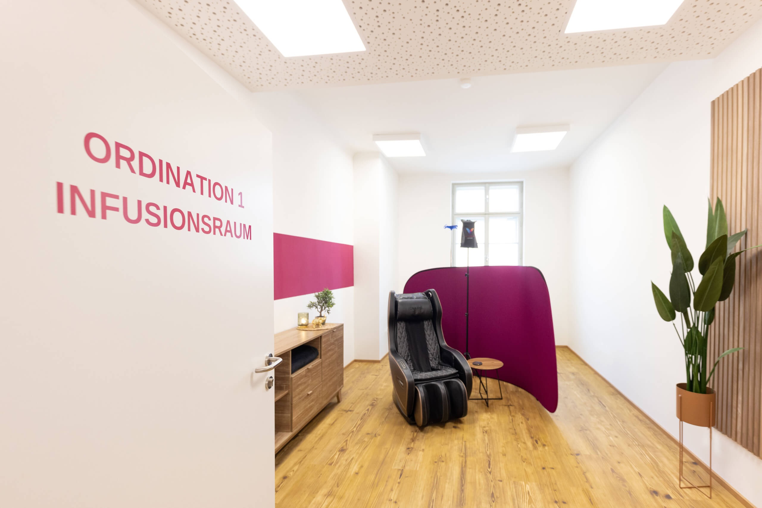 Das Bild zeigt einen modernen Infusionsraum mit einer offenen Tür, auf der "Ordination 1 Infusionsraum" in rosa Schrift steht. Der Raum ist hell und minimalistisch eingerichtet, mit Holzboden und einem gemütlichen schwarzen Massagesessel in der Mitte. Neben dem Sessel steht ein Infusionsständer, dahinter eine violette Trennwand. An der Seite ist eine kleine Holzanrichte mit Pflanzen, die dem Raum eine ruhige und einladende Atmosphäre verleihen.