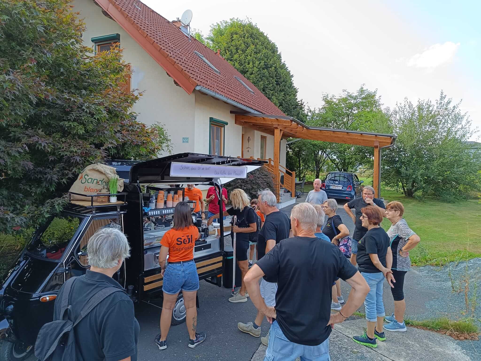 Mehrere Personen stehen um einen kleinen schwarzen Kaffeewagen, der vor einem Haus mit rotem Ziegeldach und einem Holzvordach parkt. Auf dem Kaffeewagen steht ein Schild mit der Aufschrift "Schön, dass ihr da seid! Den Kaffee des Monats probieren." Eine Frau im orangefarbenen T-Shirt und Jeans bedient die Kunden. Ein großer Sack mit der Aufschrift "Sanddorn blend" liegt auf dem Dach des Wagens. Die umstehenden Personen, meist in schwarzen T-Shirts, unterhalten sich und warten auf ihren Kaffee. Im Hintergrund sind grüne Bäume und ein Auto zu sehen. Foto: Naturfreunde Litzelsdorf