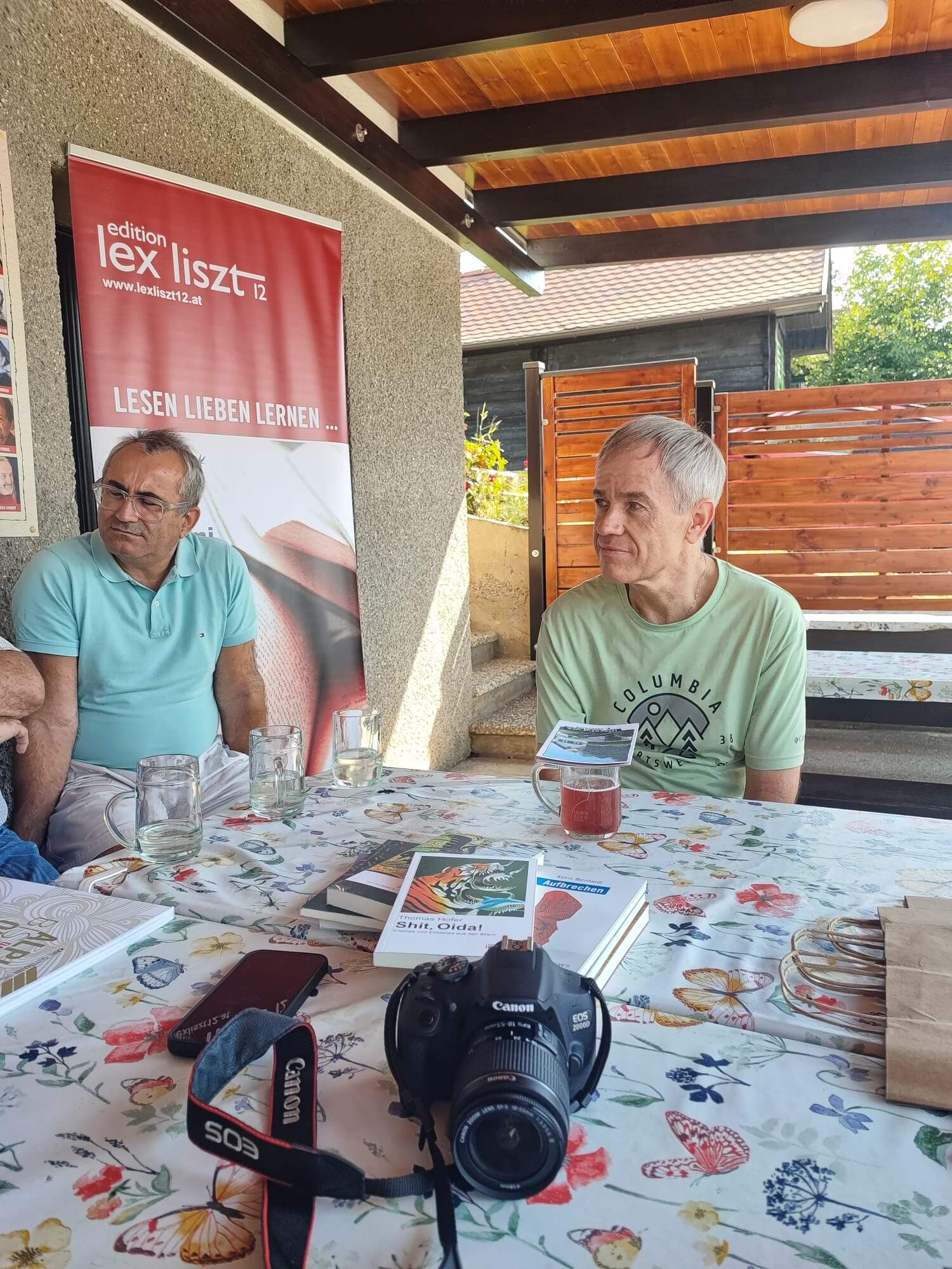 Das Bild zeigt zwei Männer, die an einem Tisch mit einer bunten, blumigen Tischdecke sitzen. Auf dem Tisch liegen Bücher, darunter eines mit dem Titel "Shit, Oida!", sowie eine Kamera und leere Trinkgläser. Im Hintergrund ist ein Banner der "edition lex liszt 12" zu sehen, auf dem der Slogan "Lesen Lieben Lernen" steht. Die Szenerie wirkt wie ein gemütliches Treffen, vermutlich in einem Außenbereich, der durch eine Überdachung und Holzwände geschützt ist. Die Atmosphäre ist entspannt und freundlich, und das Licht ist angenehm und sonnig.