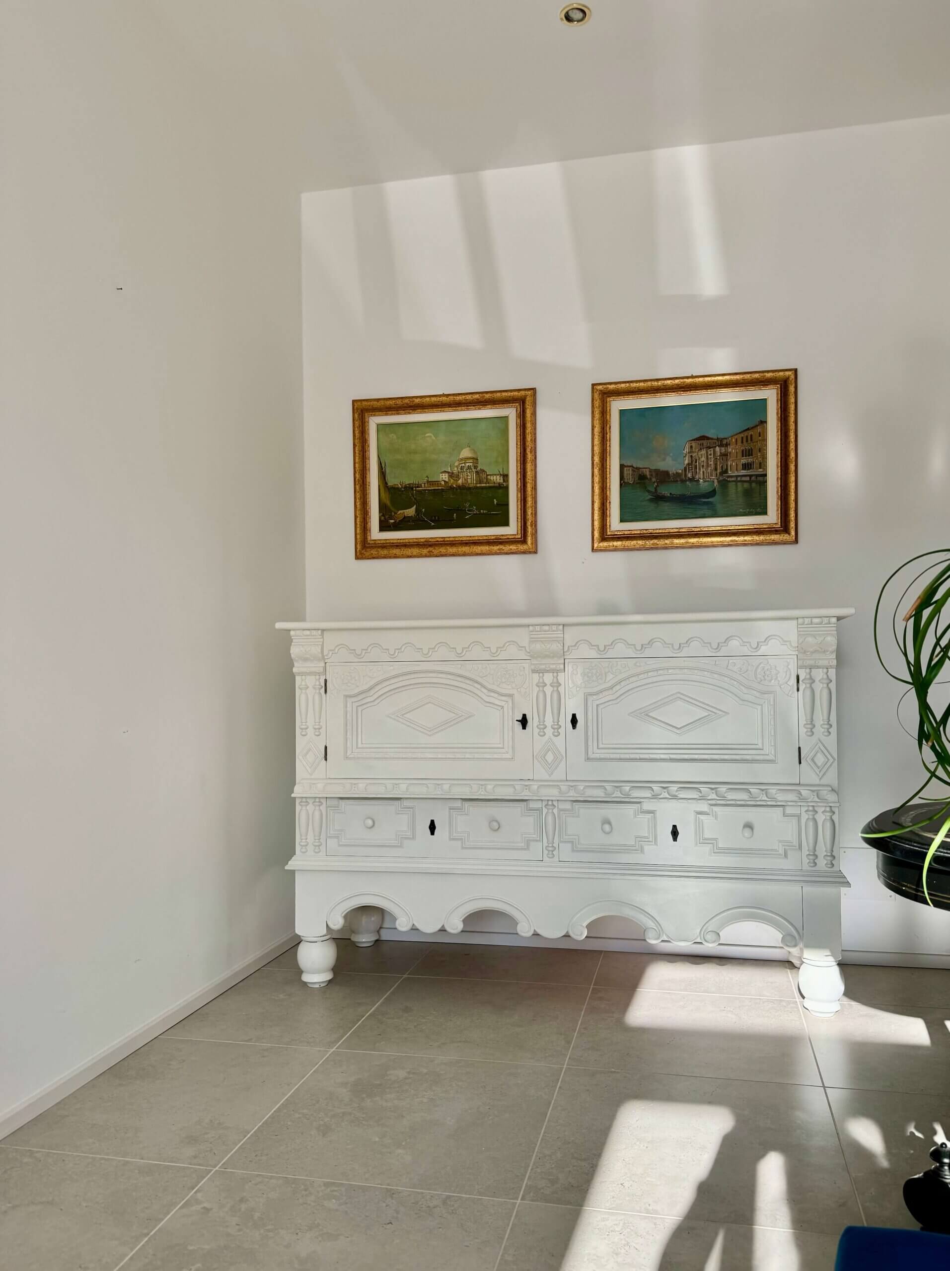 Ein weißes, reich verziertes Sideboard steht vor einer schlichten weißen Wand. Das Sideboard weist detaillierte Muster auf den Schubladen und Türen auf, mit kleinen schwarzen Schlüssellöchern. Darüber hängen zwei gerahmte Gemälde nebeneinander. Das linke Gemälde zeigt ein Kuppelgebäude an einem Wasserufer, während das rechte eine Kanalszene mit Gebäuden am Wasser und einem Boot darstellt. Beide Gemälde haben goldfarbene Rahmen. Rechts im Bild ist eine grüne Pflanze in einem schwarzen Topf teilweise sichtbar. Sonnenlicht fällt herein und wirft weiche Schatten auf den gefliesten Boden und die Wand. Foto Kasumovic