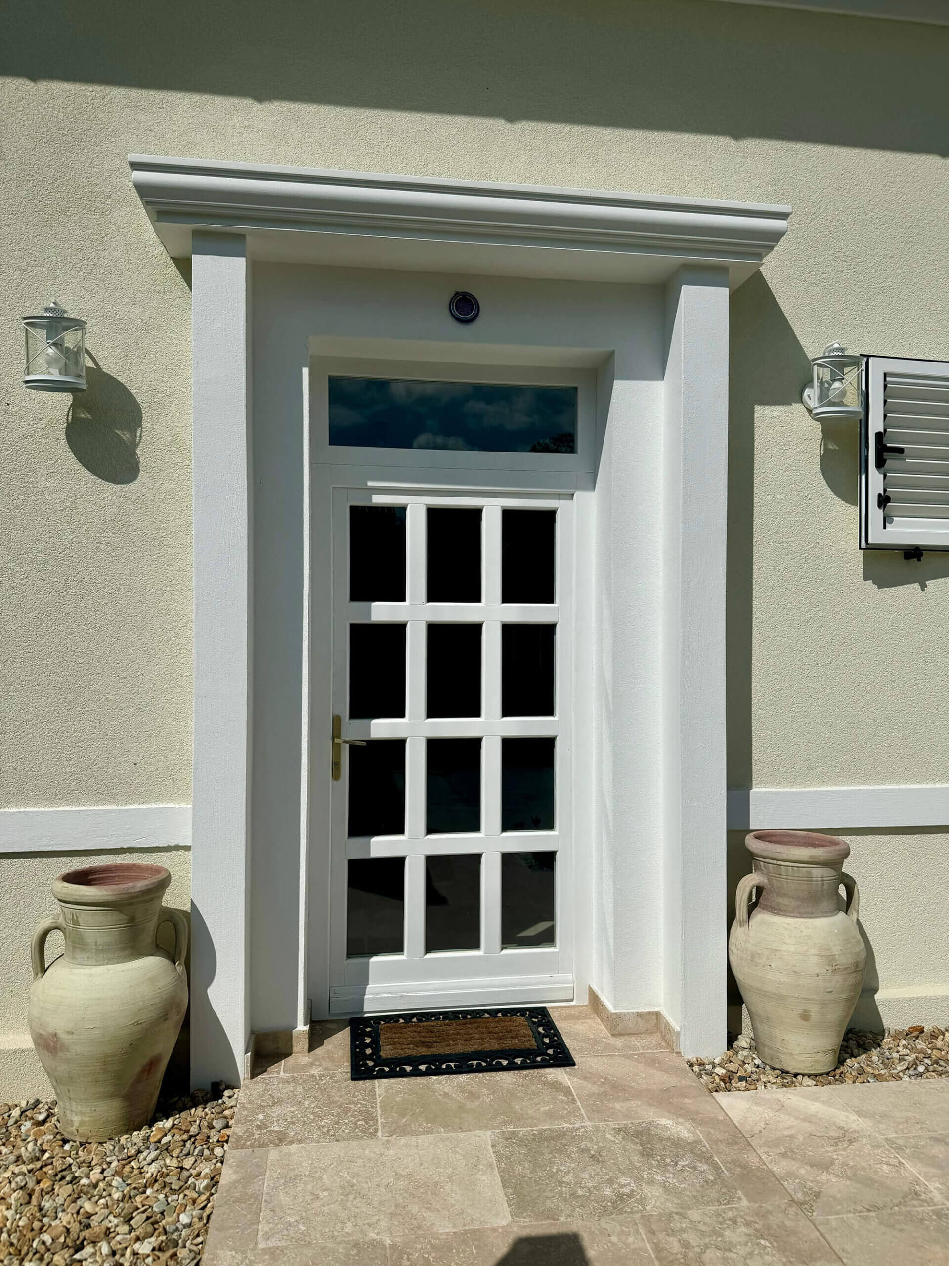 Eine weiße Eingangstür mit Glasfenstern, die in einem symmetrischen Gittermuster angeordnet sind, ist zentral in der hellbeigen Fassade eines Hauses eingelassen. Die Tür ist von einem weißen Rahmen mit einem kleinen Vordach umgeben. Links und rechts neben der Tür stehen große, beigefarbene Amphoren aus Terrakotta auf einem mit Kieselsteinen bedeckten Streifen. Zwei klassische Wandlaternen in Metall- und Glasoptik sind jeweils links und rechts oberhalb der Amphoren angebracht. Vor der Tür liegt eine Fußmatte auf dem gefliesten Eingangsbereich..Foto: Kasumovic
