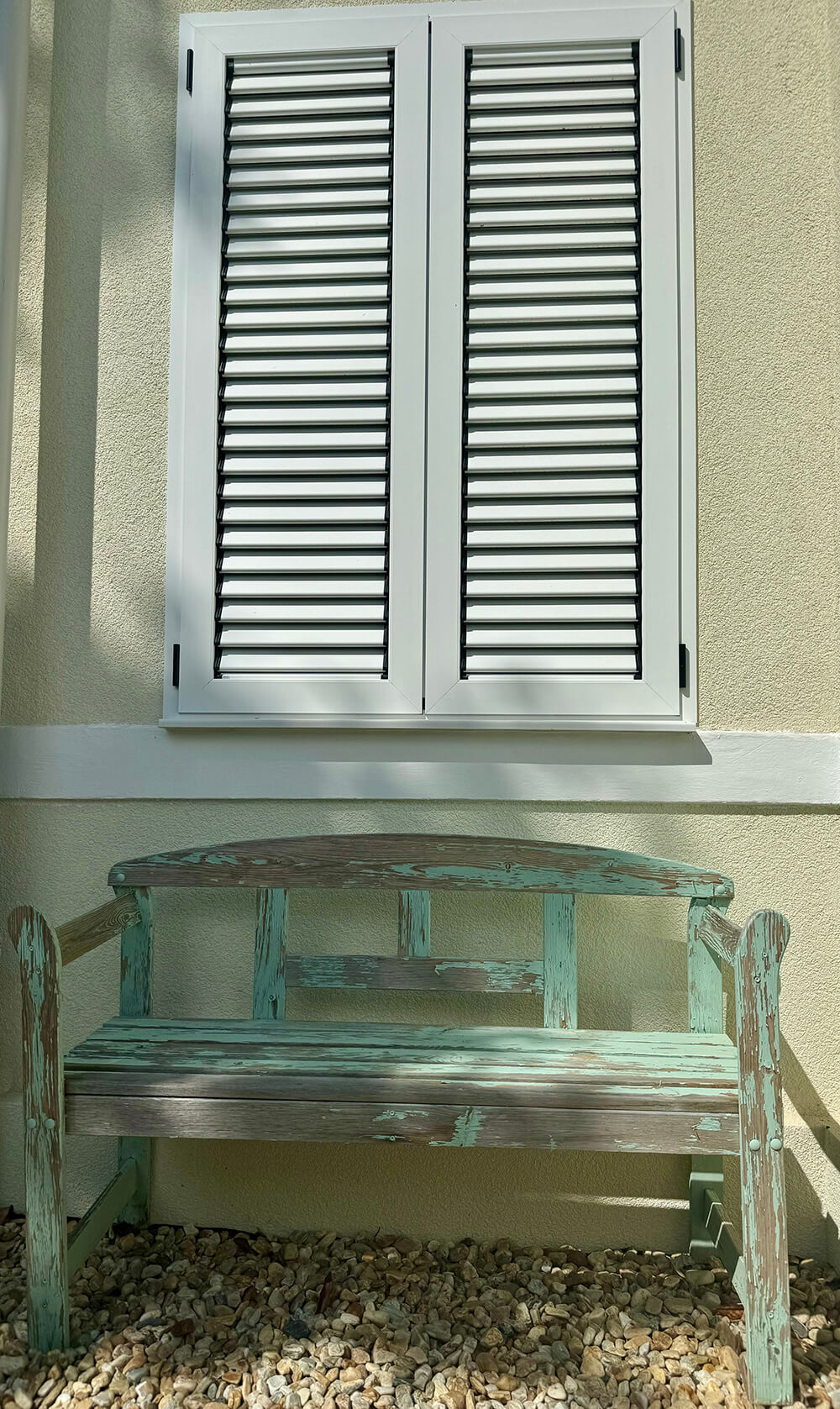Eine verwitterte Holzbank mit abblätternder türkisfarbener Farbe steht vor einer hellbeigen Hauswand. Über der Bank befindet sich ein geschlossenes Fenster mit weißen Lamellenläden. Der Boden unter der Bank ist mit kleinen, hellen Kieselsteinen bedeckt. Die Szenerie wird von Schatten durchzogen, die auf die Wand und die Bank fallen, was auf Sonnenschein und umliegende Bäume hindeutet. Foto:Kasumovic
