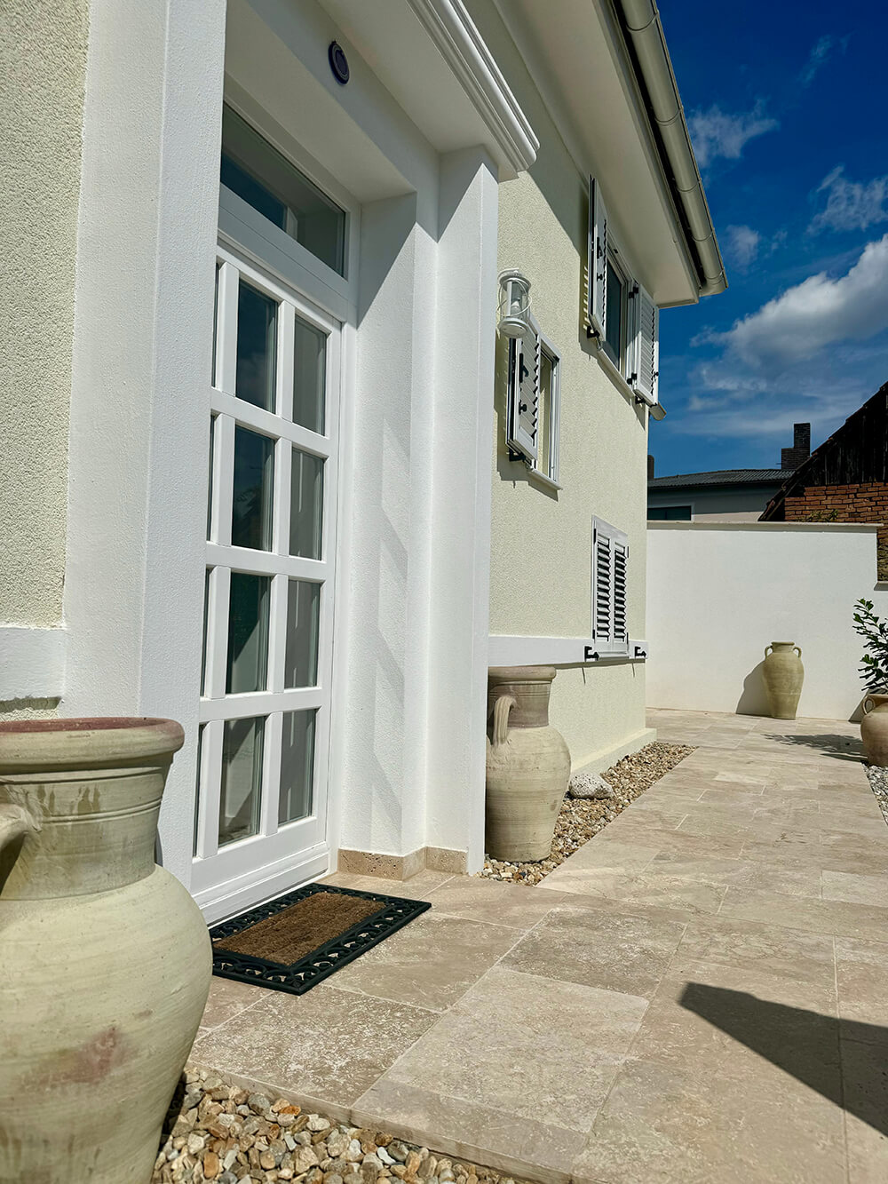 Ein Eingang mit einer weißen Tür, die in einem hellbeigen Haus eingelassen ist. Die Tür hat ein Glasgitter und wird von einem weißen Rahmen mit einem kleinen Vordach umgeben. Vor der Tür liegt eine Fußmatte auf einem gefliesten Boden. Links und rechts neben der Tür stehen große, beigefarbene Terrakotta-Amphoren auf einem mit Kieselsteinen bedeckten Streifen. Entlang des Hauses sind weitere Amphoren platziert. An der Fassade ist eine Laterne aus Metall und Glas angebracht. Der Himmel ist klar und blau, und die Sonne wirft Schatten auf den Boden. Foto: Kasumovic