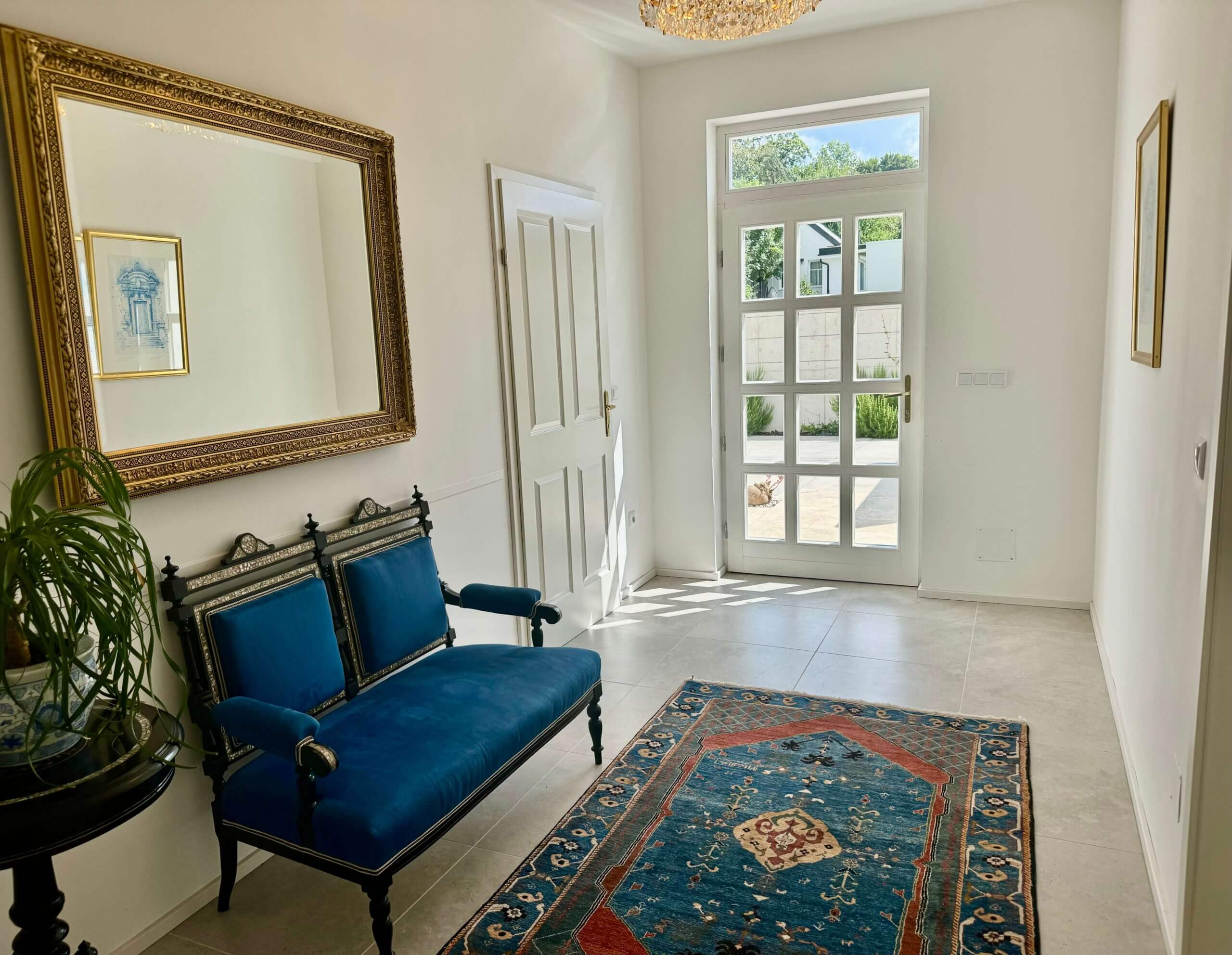 Ein heller Flur mit weißen Wänden und hellen Fliesen. An der linken Wand hängt ein großer, reich verzierter Spiegel mit einem goldfarbenen Rahmen. Darunter steht ein antikes Sofa mit schwarzem Holzrahmen und blauen Polstern. Daneben ist ein kleiner runder Tisch mit einer grünen Pflanze darauf zu sehen. Auf dem Boden liegt ein großer, gemusterter Teppich in Blau-, Rot- und Beigetönen. Rechts im Bild befindet sich eine weiße Tür mit Glasfenstern, die nach außen führt und durch die man einen Garten mit grünen Pflanzen sehen kann. An der rechten Wand hängt ein kleines, gerahmtes Bild. Foto:Kasumovic