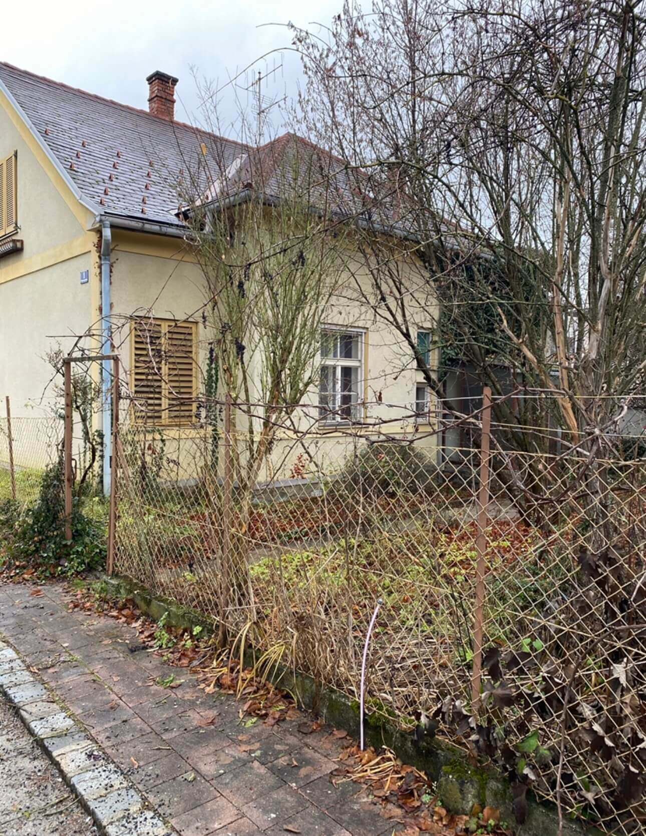 Ein älteres, beigefarbenes Haus mit einem grauen Schrägdach und einem roten Schornstein. Die Fassade zeigt zwei Fenster, eines mit braunen Fensterläden. Vor dem Haus steht ein Metallzaun mit Maschendraht, der ein verwildertes Gartenstück umgibt. Im Garten sind kahle Bäume und Sträucher zu sehen, deren Äste teils über den Zaun ragen. Der Gehweg vor dem Zaun ist mit braunen und grauen Pflastersteinen belegt und von herabgefallenen Blättern bedeckt. Der Himmel ist bewölkt, was auf ein kühles, trübes Wetter hinweist. Foto:Kasumovic