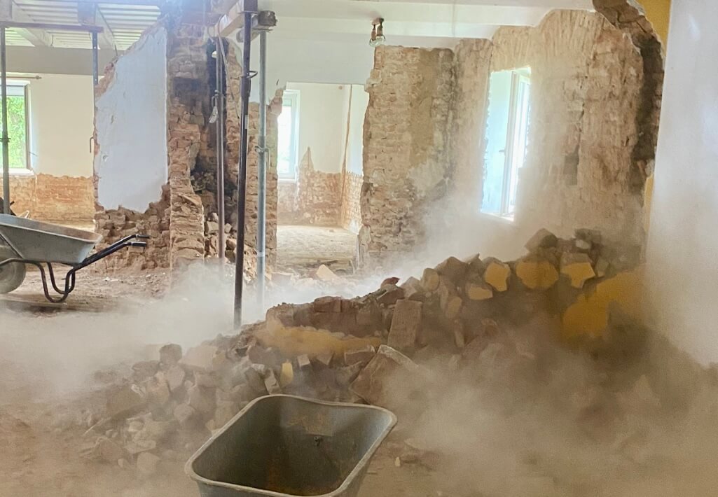 Eine Baustelle im Inneren eines Gebäudes zeigt teilweise abgerissene Wände, freigelegtes Mauerwerk und herumliegende Trümmer. Dicker Staub schwebt in der Luft, teilweise verdeckt er den Boden. Im Vordergrund steht eine Schubkarre, daneben ein leeres Bauwännchen. Metallstützen halten die Decke, während zwei Fenster im Hintergrund schwaches Tageslicht hereinlassen. Die Szene vermittelt den Eindruck von Abrissarbeiten oder umfangreicher Renovierung. Foto: Kasumovic