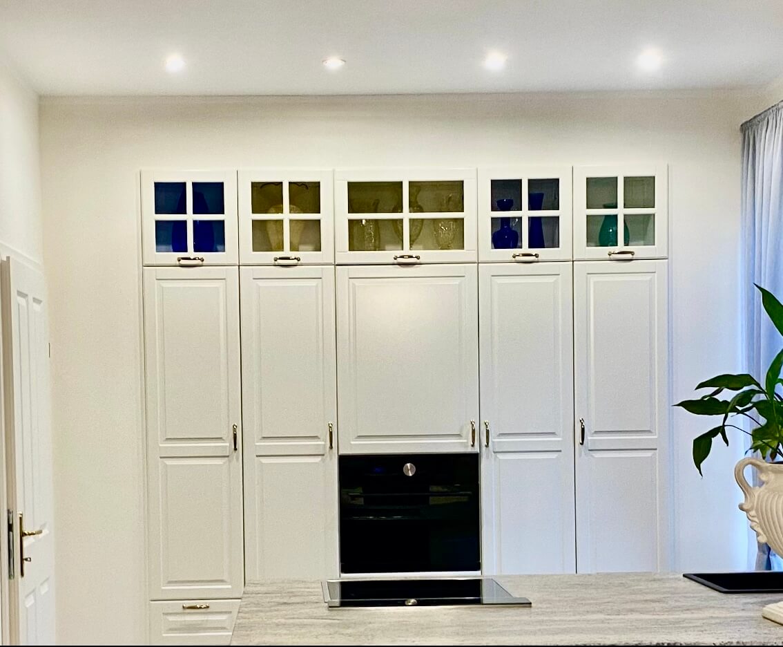 Eine moderne Küche mit einer Wand aus hohen, weißen Schränken, die bis zur Decke reichen. Im mittleren Bereich ist ein eingebauter Backofen zu sehen. Über den Schränken befinden sich kleine Glastüren mit darin platzierten Vasen und Gläsern in Blau- und Grüntönen. Die Arbeitsfläche im Vordergrund besteht aus hellem Stein. Rechts im Bild steht eine große Zimmerpflanze neben einem bodenlangen, weißen Vorhang. Die Decke ist mit mehreren kleinen, eingebauten Strahlern beleuchtet. Foto: Kasumovic