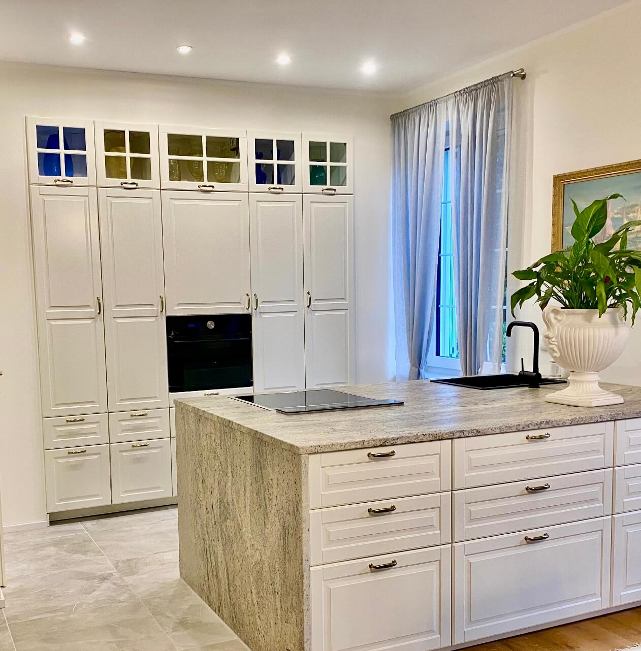 Eine moderne Küche mit einer Wand aus hohen, weißen Schränken, die bis zur Decke reichen. Im mittleren Bereich ist ein eingebauter Backofen zu sehen. Über den Schränken befinden sich kleine Glastüren mit darin platzierten Vasen und Gläsern in Blau- und Grüntönen. Die Arbeitsfläche im Vordergrund besteht aus hellem Stein. Rechts im Bild steht eine große Zimmerpflanze neben einem bodenlangen, weißen Vorhang. Die Decke ist mit mehreren kleinen, eingebauten Strahlern beleuchtet. Foto: Kasumovic