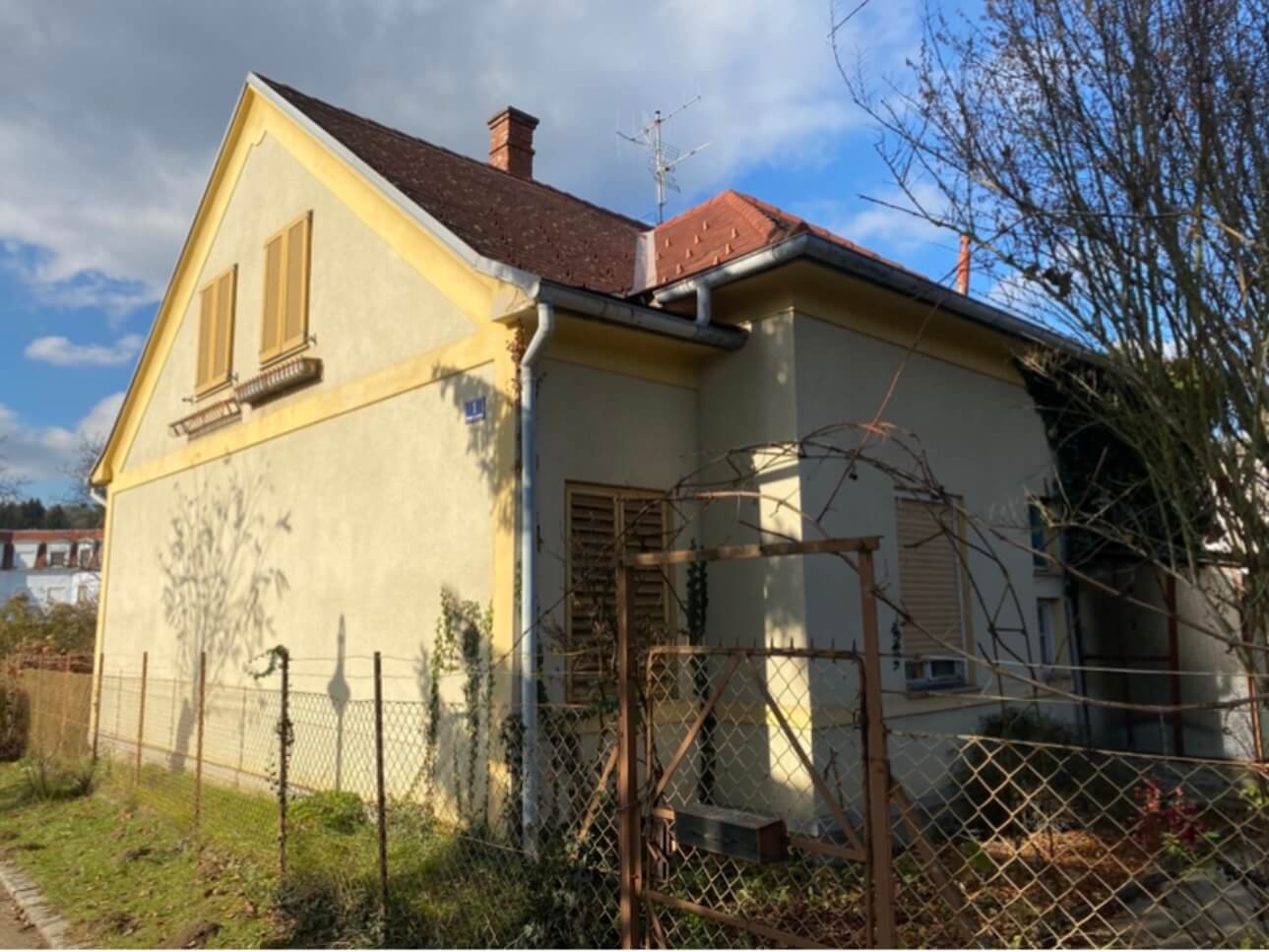Ein kleines, älteres Haus mit hellgelber Fassade und rotbraunem Satteldach. Die Fensterläden sind geschlossen, und am oberen Fenster befinden sich Blumenkästen. Eine einfache, metallene Regenrinne verläuft an der Ecke des Hauses. Vor dem Haus steht ein metallener Maschendrahtzaun mit einem schmalen Tor. Eine Pergola ohne Blätter ist über dem Torbogen sichtbar. Ein kahler Baum steht rechts im Bild, und einige Kletterpflanzen ranken am Zaun empor. Im Hintergrund sind einige Gebäude und Bäume unter einem teilweise bewölkten Himmel zu sehen. Foto: KAsumovic