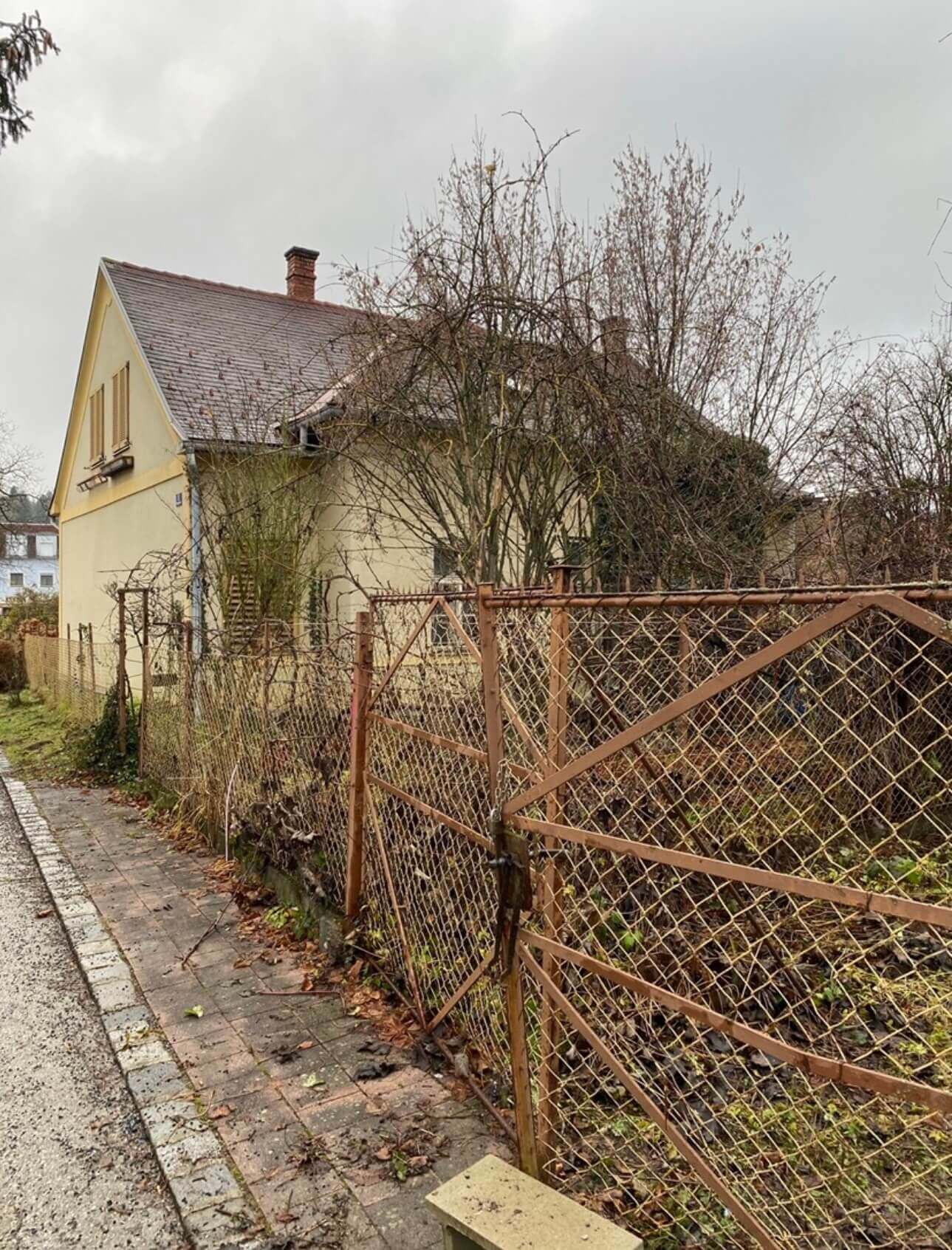Ein beiges, älteres Haus mit grauem Schrägdach und einem roten Schornstein. Das Haus hat zwei sichtbare Fenster, eines davon mit braunen Fensterläden. Vor dem Haus verläuft ein Metallzaun mit Maschendraht, der einen verwilderten Garten abgrenzt. Im Garten stehen kahle Bäume und Sträucher, deren Äste über den Zaun ragen. Der Gehweg vor dem Zaun ist mit Pflastersteinen belegt und von herabgefallenen Blättern bedeckt. Es ist bewölkt, was auf trübes und kühles Wetter hinweist. Foto:Kasumovic