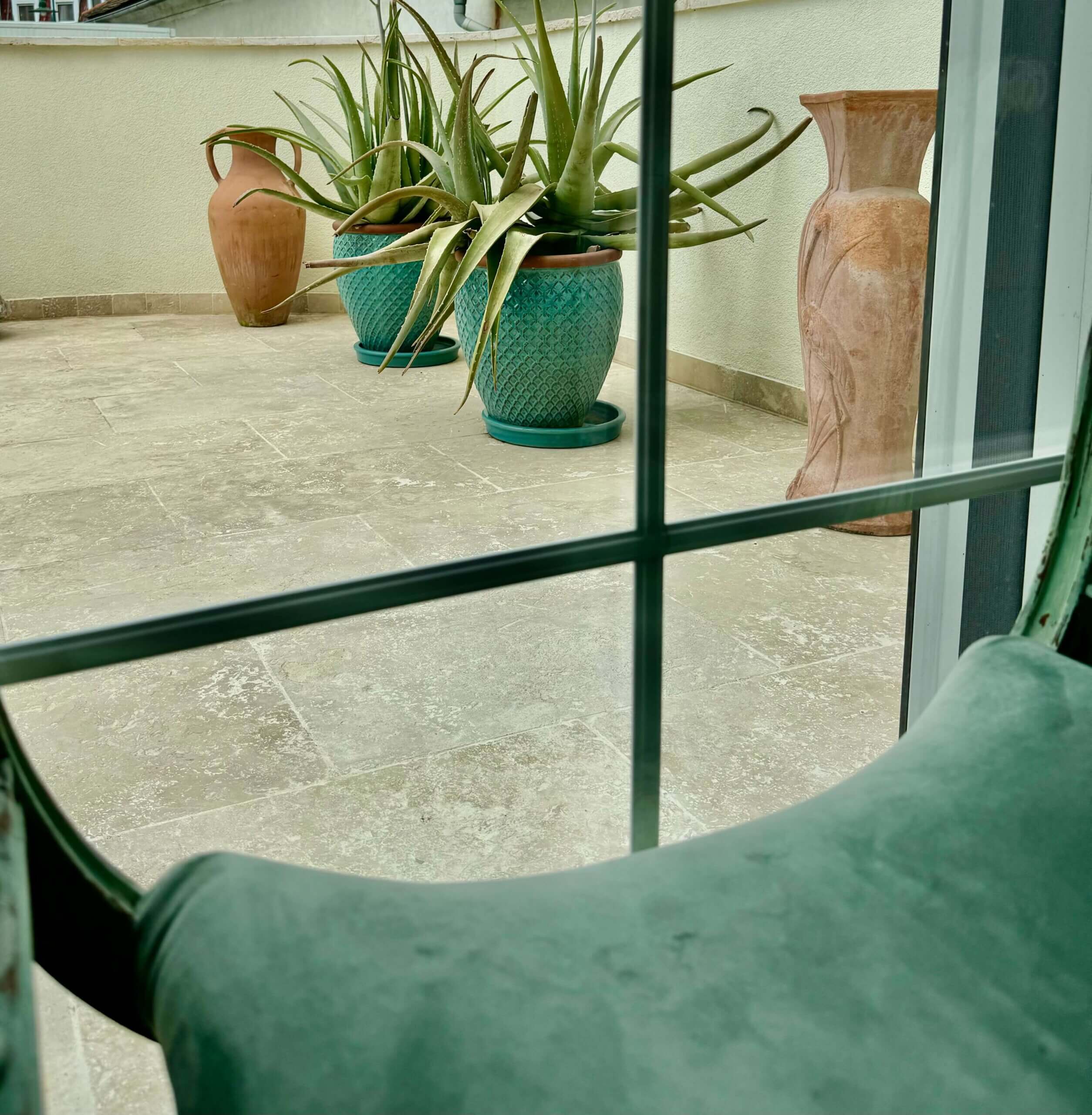 Eine Terrasse mit hellen Steinfliesen, auf der drei große Pflanzgefäße stehen. Zwei grüne, strukturierte Töpfe enthalten große, spitz zulaufende Sukkulenten, während die beiden anderen Töpfe terrakottafarben und bauchig geformt sind und leer wirken. Die Terrasse ist von einer cremefarbenen Mauer umgeben. Im Vordergrund des Bildes ist ein Teil eines grünen Polsterstuhls durch ein Fenster sichtbar, das durch Sprossen in vier Abschnitte unterteilt ist. Foto: Kasumovic