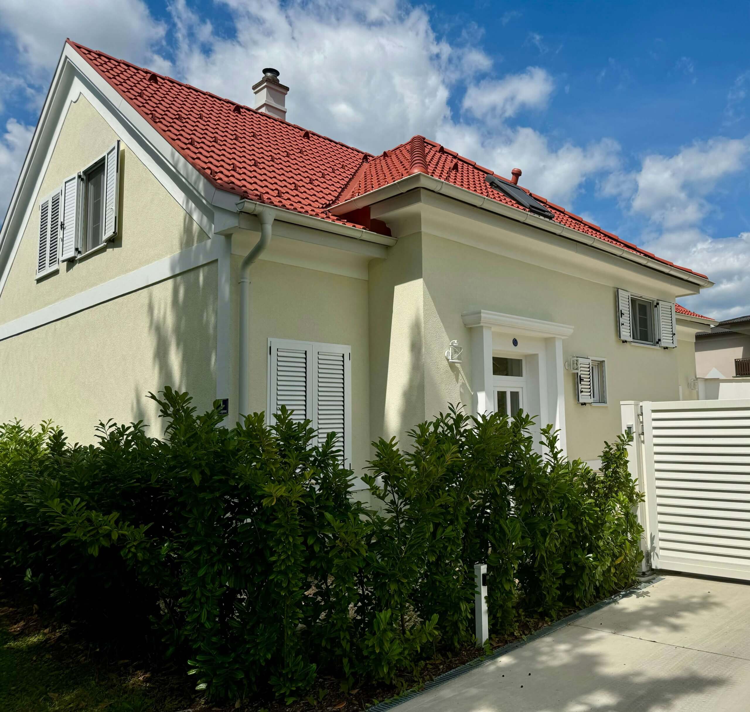 Ein modernes, hellbeiges Haus mit roten Dachziegeln und weißen Fensterläden. Das Dach hat mehrere Schrägen, darunter einen kleinen Schornstein und ein Dachfenster. Die Fassade ist schlicht gestaltet, mit einer weißen Eingangstür, die von einem kleinen Vordach geschützt wird. Links neben der Tür stehen dichte, grüne Sträucher, die bis unter die Fenster reichen. Rechts im Bild ist ein weißes Lamellen-Gartentor zu sehen, das zur gepflasterten Einfahrt führt. Der Himmel ist blau mit vereinzelten weißen Wolken. Foto: Kasumovic