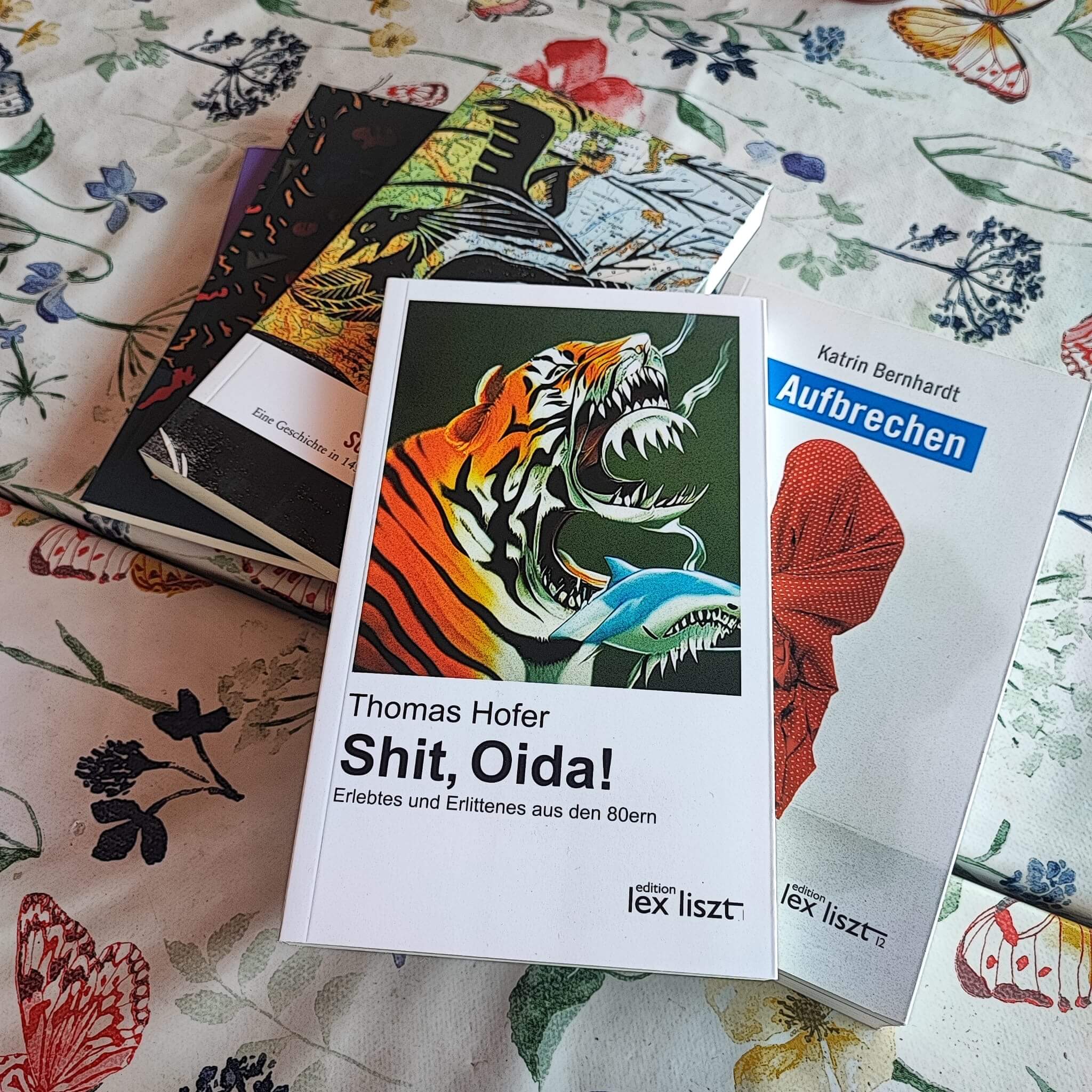 Das Bild zeigt eine Nahaufnahme von vier Büchern, die leicht übereinander auf einer blumigen Tischdecke liegen. Im Vordergrund ist das Buch *"Shit, Oida!"* von Thomas Hofer zu sehen, dessen Cover eine farbenfrohe Illustration eines brüllenden Tigers und eines Haifisches zeigt. Daneben liegt das Buch *"Aufbrechen"* von Katrin Bernhardt, das ein rotes, gefaltetes Kleidungsstück auf dem Cover darstellt. Im Hintergrund sind zwei weitere Bücher mit kunstvollen und detailreichen Covern zu erkennen. Die Szene vermittelt eine lebendige literarische Atmosphäre.