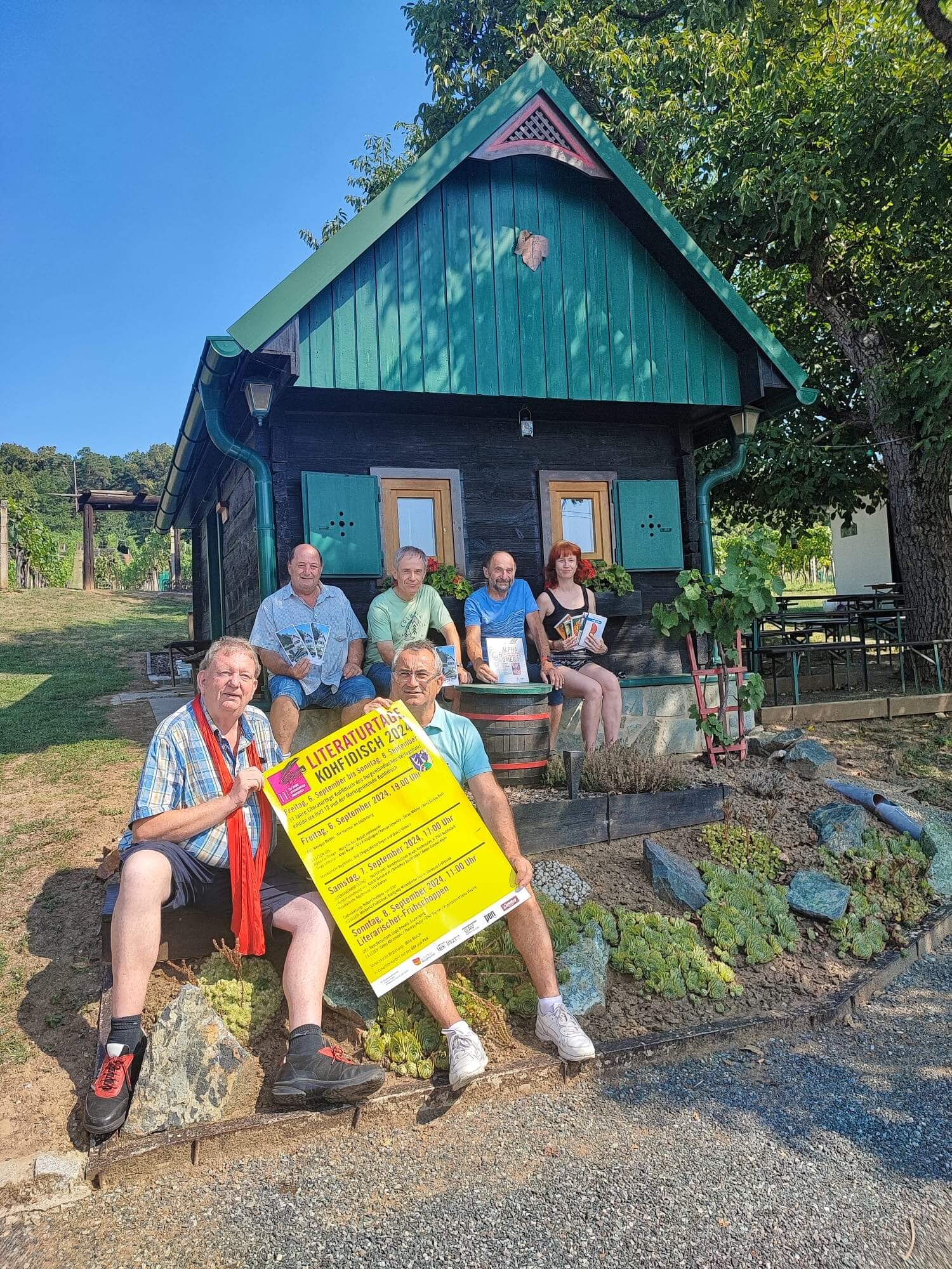 Das Bild zeigt sechs Personen, die auf einer Steinmauer vor einer kleinen, traditionellen Holzhütte mit grünen Fensterläden und einem grünen Dach sitzen. Zwei der Personen halten ein großes, gelbes Plakat der "Literaturtage Kohfidisch 2024" in den Händen. Die anderen halten Bücher. Die Umgebung ist ländlich, mit einem Baum rechts im Bild und grüner Vegetation im Hintergrund. Das Wetter ist sonnig, und die Atmosphäre wirkt freundlich und entspannt. Die Personen tragen legere Kleidung und sitzen nah beieinander, was eine gewisse Verbundenheit vermittelt.
