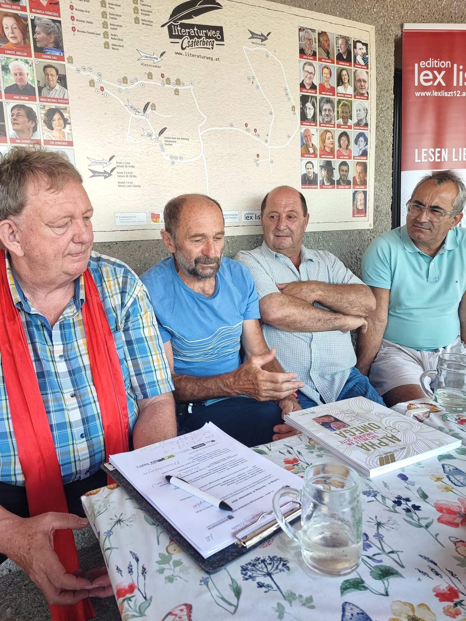 Das Bild zeigt vier Männer, die eng nebeneinander an einem Tisch mit einer blumigen Tischdecke sitzen. Vor ihnen liegen Notizunterlagen und Bücher, unter anderem ein Buch mit dem Titel "Teta Mare". Einer der Männer spricht gestikulierend, während die anderen aufmerksam zuhören. Im Hintergrund befindet sich eine Karte des "Literaturwegs Csaterberg" sowie ein Poster mit Fotos von Autor*innen. Rechts ist erneut das Banner der "edition lex liszt 12" zu sehen, auf dem der Slogan "Lesen Lieben Lernen" steht. Die Atmosphäre wirkt wie eine konzentrierte Gesprächsrunde, möglicherweise im Rahmen eines literarischen Events oder Treffens.