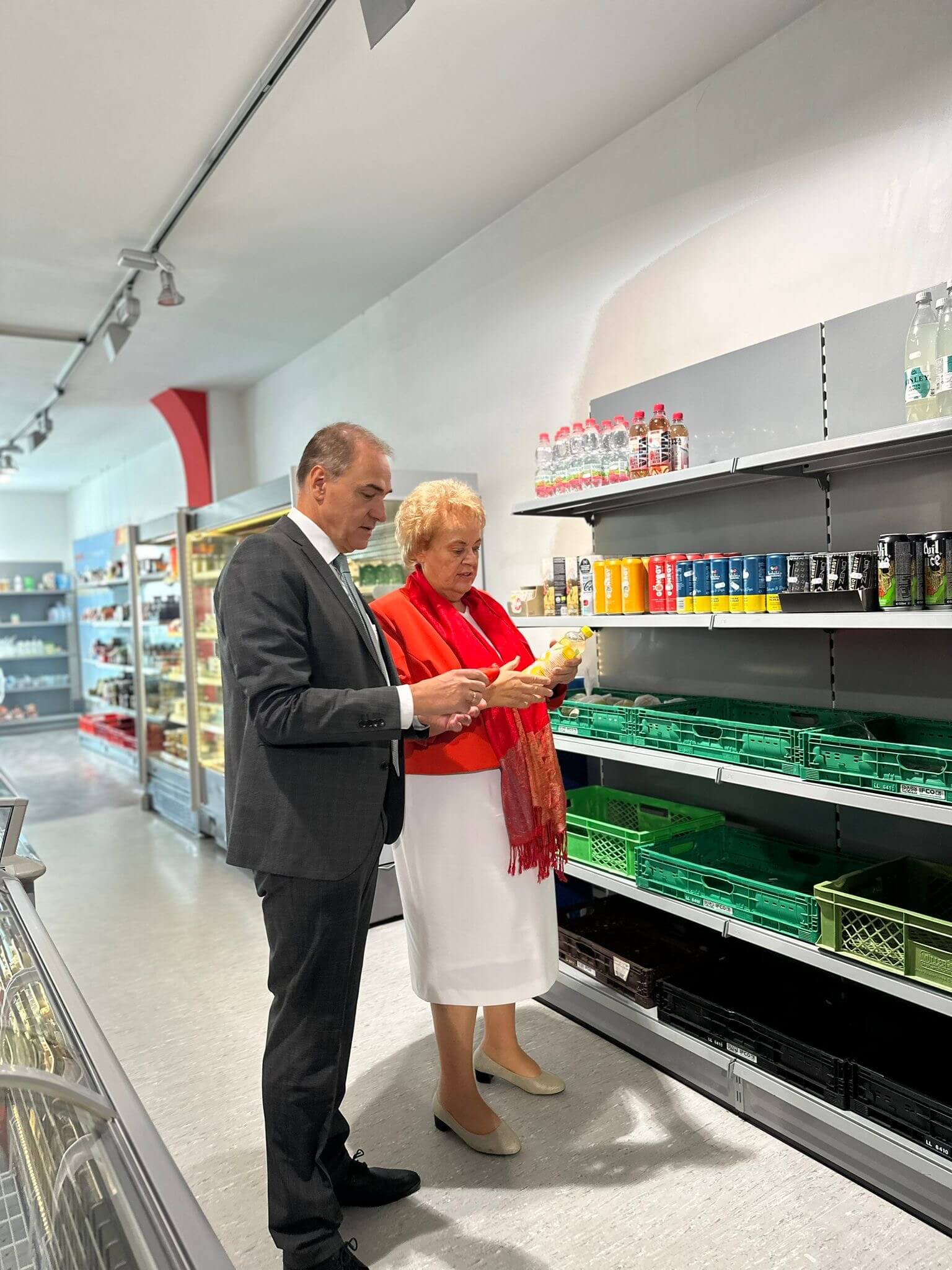 Leonhard Schneemann und Verena Dunst stehen gemeinsam vor einem Regal im Sonnenmarkt. Beide halten ein Produkt in den Händen und scheinen sich darüber auszutauschen. Verena Dunst trägt einen roten Blazer und einen weißen Rock, Leonhard Schneemann ist in einem dunklen Anzug gekleidet. Im Hintergrund sind Regale mit Getränken und Lebensmitteln zu sehen.