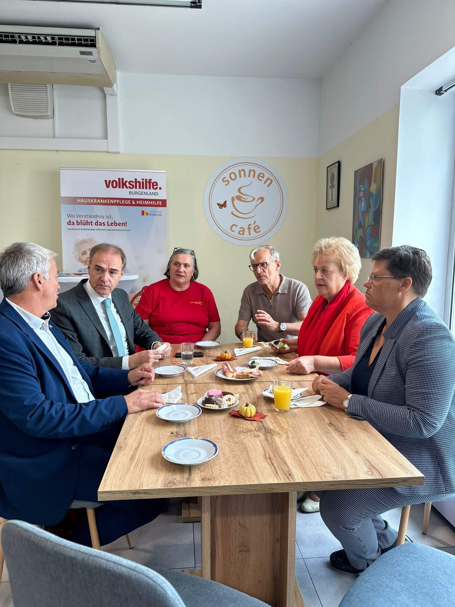  Drei Personen stehen im Sonnenmarkt vor einem Regal mit Lebensmitteln. Links im Bild ist Leonhard Schneemann in einem grauen Anzug zu sehen, in der Mitte steht Sylvia Hajszan, die Marktleiterin, in einem roten T-Shirt. Rechts steht Verena Dunst, gekleidet in einem roten Blazer und einem weißen Rock. Alle drei halten Broschüren des Sonnenmarkts in den Händen und lächeln in die Kamera. Im Hintergrund sind Regale mit Produkten sowie ein Banner mit den Logos der Volkshilfe und des Landes Burgenland zu sehen.