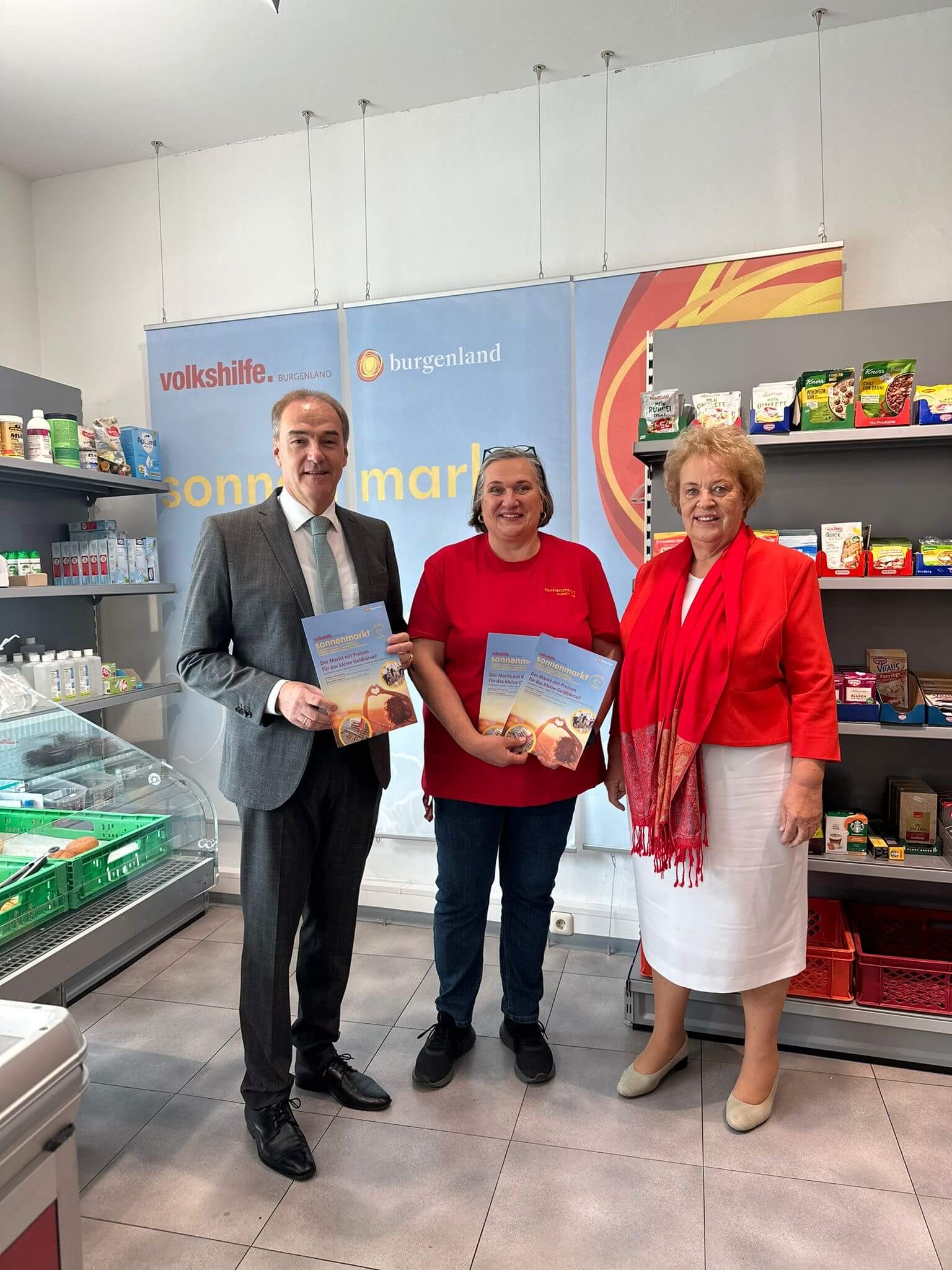 Sechs Personen sitzen gemeinsam an einem Tisch im Sonnencafé und führen ein Gespräch. Es sind von links nach rechts ein Mann im dunklen Anzug, Leonhard Schneemann, Sylvia Hajszan im roten T-Shirt, ein Mann in grauem Polo-Shirt, Verena Dunst in rotem Blazer und weißem Rock, sowie eine Frau im grauen Blazer. Auf dem Tisch befinden sich Getränke, kleine Speisen und Gebäck. Im Hintergrund sind ein Banner der Volkshilfe Burgenland und das Logo des Sonnencafés an der Wand zu sehen. Die Atmosphäre wirkt freundlich und entspannt.