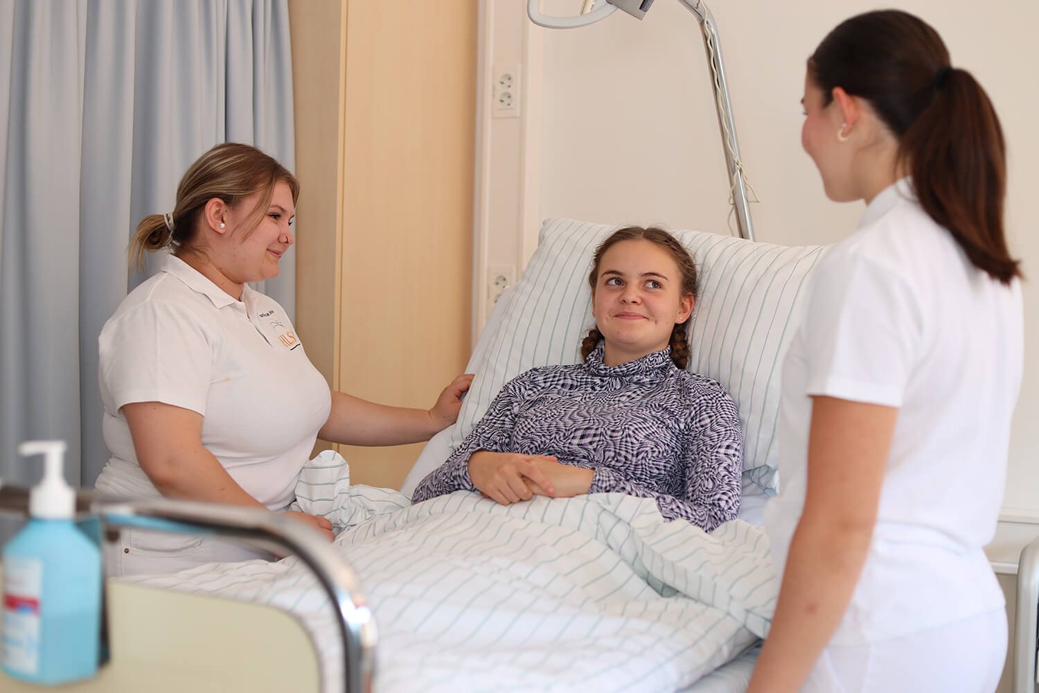 Eine junge Patientin liegt in einem Krankenhausbett, zugedeckt mit einer weiß-blau gestreiften Decke. Sie trägt ein langärmeliges Oberteil mit einem violett-weißen Muster und lächelt leicht, während sie zwei Pflegekräfte anschaut. Beide Pflegekräfte tragen weiße Kleidung, die linke Person steht näher am Bett und hält eine Hand auf die Bettdecke, während die rechte Pflegekraft am Fußende des Bettes steht. Im Vordergrund ist ein Spender für Desinfektionsmittel zu sehen. Der Raum wirkt hell und freundlich. Foto: HLW Pinkafeld