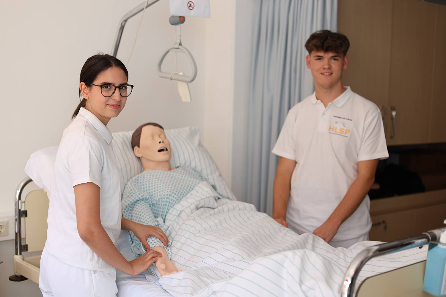 Zwei Pflegeschüler, eine Frau links mit Brille und ein junger Mann rechts, stehen an einem Krankenhausbett mit einer Krankenpflegepuppe. Beide tragen weiße Kleidung. Die Frau berührt den Arm der Puppe, die mit einer weiß-blau gestreiften Decke zugedeckt ist und ein Krankenhaushemd trägt. Über dem Bett hängt ein Haltegriff. Im Hintergrund sind hellblaue Vorhänge und ein Schrank zu sehen. Der Raum wirkt klinisch und hell, geeignet für Pflege- oder medizinische Übungszwecke. Foto: HLW Pinkafeld