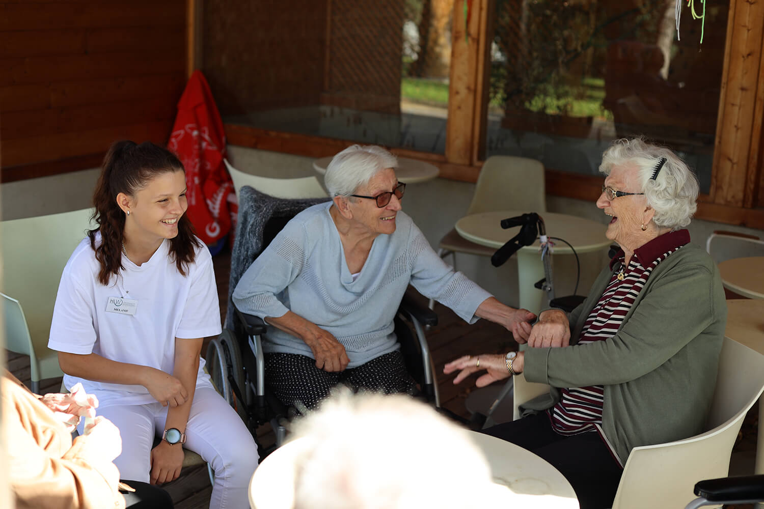 Drei Frauen sitzen an einem runden Tisch in einer gemütlichen, überdachten Außenumgebung. Links sitzt eine junge Pflegerin in weißer Kleidung mit Namensschild und lächelt. In der Mitte sitzt eine ältere Frau im Rollstuhl, die eine hellgraue Strickjacke trägt und ebenfalls lächelt. Sie hält die Hand der älteren Frau rechts, die eine grünliche Strickjacke über einem rot-weiß gestreiften Poloshirt trägt und lacht herzlich. Im Hintergrund sind Stühle, Tische und eine Fensterfront zu sehen, draußen scheint die Sonne. Eine Gehhilfe steht neben dem Tisch. Foto: HLW Pinkafeld