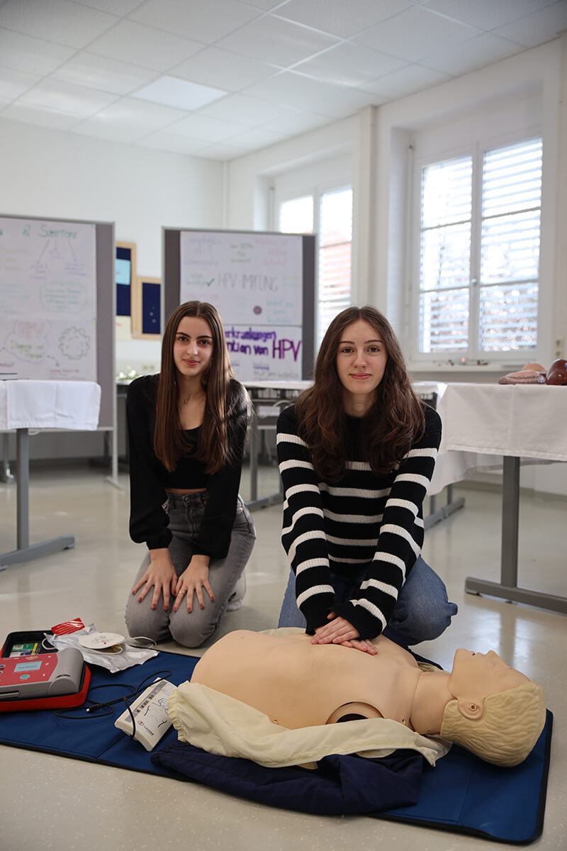 Zwei junge Frauen knien in einem Schulungsraum auf dem Boden neben einer Reanimationspuppe. Die Frau links trägt ein schwarzes Oberteil, während die Frau rechts ein schwarz-weiß gestreiftes Oberteil trägt und mit beiden Händen eine Herzdruckmassage an der Puppe durchführt. Neben der Puppe liegt ein Defibrillator und Zubehör. Im Hintergrund sind Poster und Schautafeln mit Informationen zu medizinischen Themen, darunter „HPV“, zu sehen. Der Raum ist hell, mit großen Fenstern und Tischen. Foto: HLW Pinkafeld