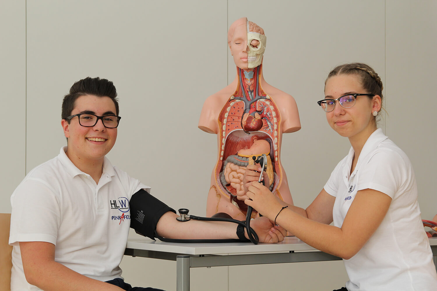 Ein junger Mann und eine junge Frau sitzen an einem Tisch, beide tragen weiße Polohemden mit dem Logo „HLW Pinkafeld“. Der junge Mann lächelt und hat eine Blutdruckmanschette am linken Arm angelegt, während die junge Frau ihm mit einem Stethoskop den Blutdruck misst. Hinter ihnen steht ein anatomisches Modell eines menschlichen Oberkörpers mit offenen Organen, das zur Veranschaulichung dient. Beide tragen Brillen und der Raum ist schlicht und hell gestaltet. Foto: HLW Pinkafeld
