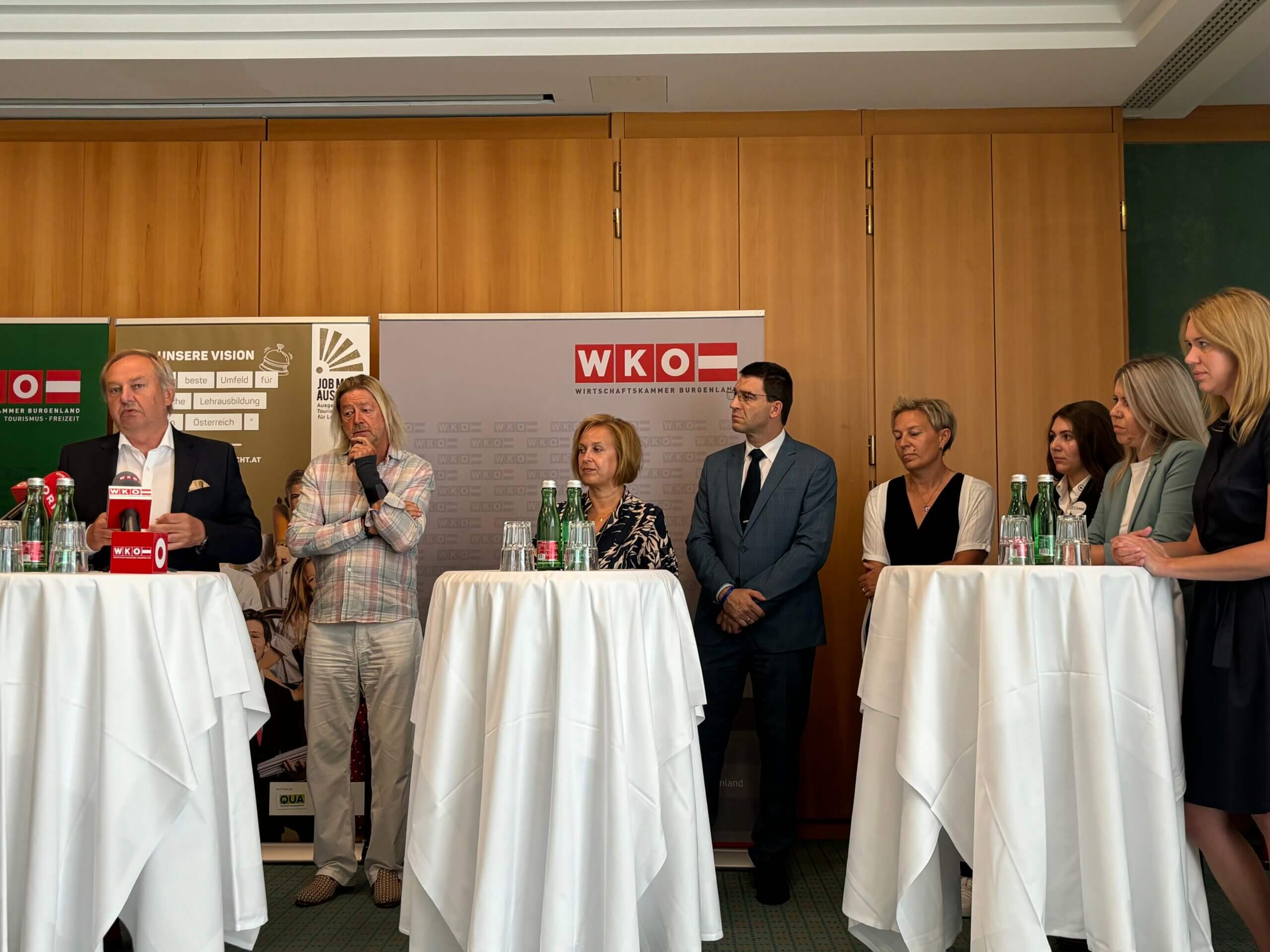 Eine Gruppe von sieben Personen steht oder sitzt auf einer Bühne. Im Mittelpunkt spricht ein Mann hinter einem Tisch mit Mikrofonen. Im Hintergrund sind Banner der WKO und das Logo von "JOB MIT AUSSICHT" sichtbar.