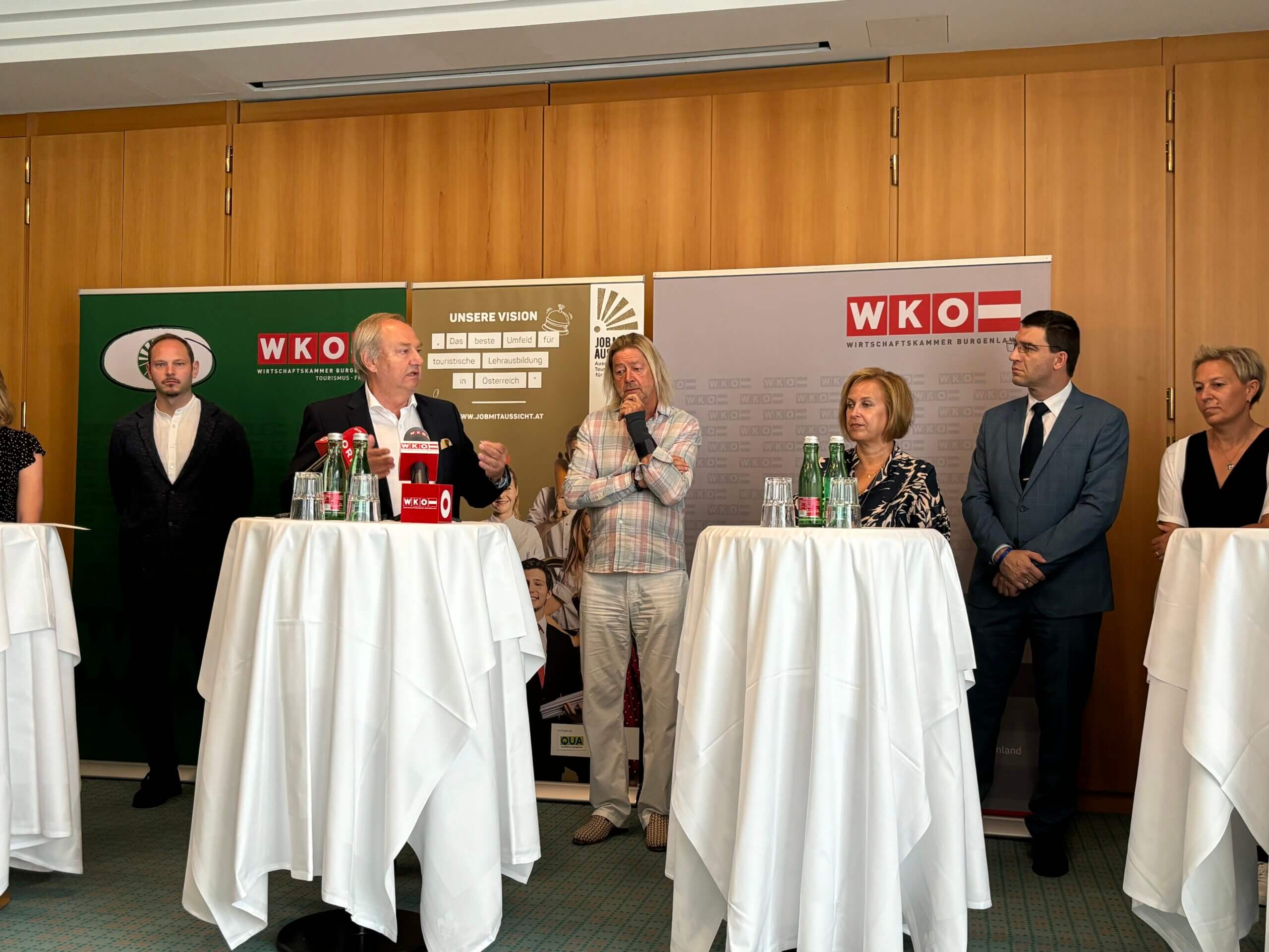 Mehrere Personen stehen auf einer Bühne und sprechen in Mikrofone. Im Hintergrund sind Banner der WKO zu sehen, und mehrere Teilnehmer hören aufmerksam zu.