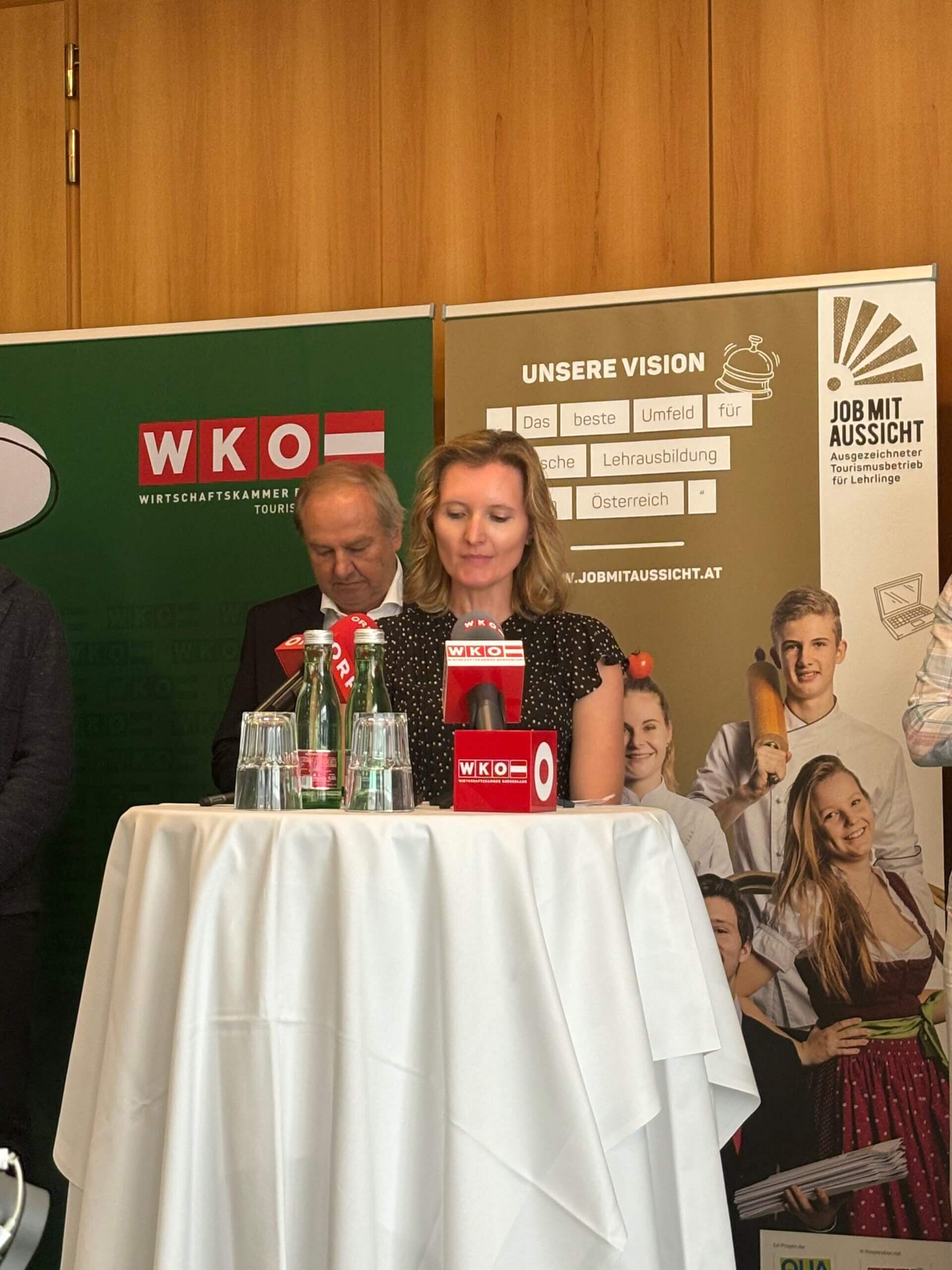 Marlies Rainer steht hinter einem Tisch und spricht in Mikrofone. Vor ihr stehen mehrere Mikrofone mit dem Logo der Wirtschaftskammer Österreich (WKO) auf einem Tisch mit weißen Tischdecken.