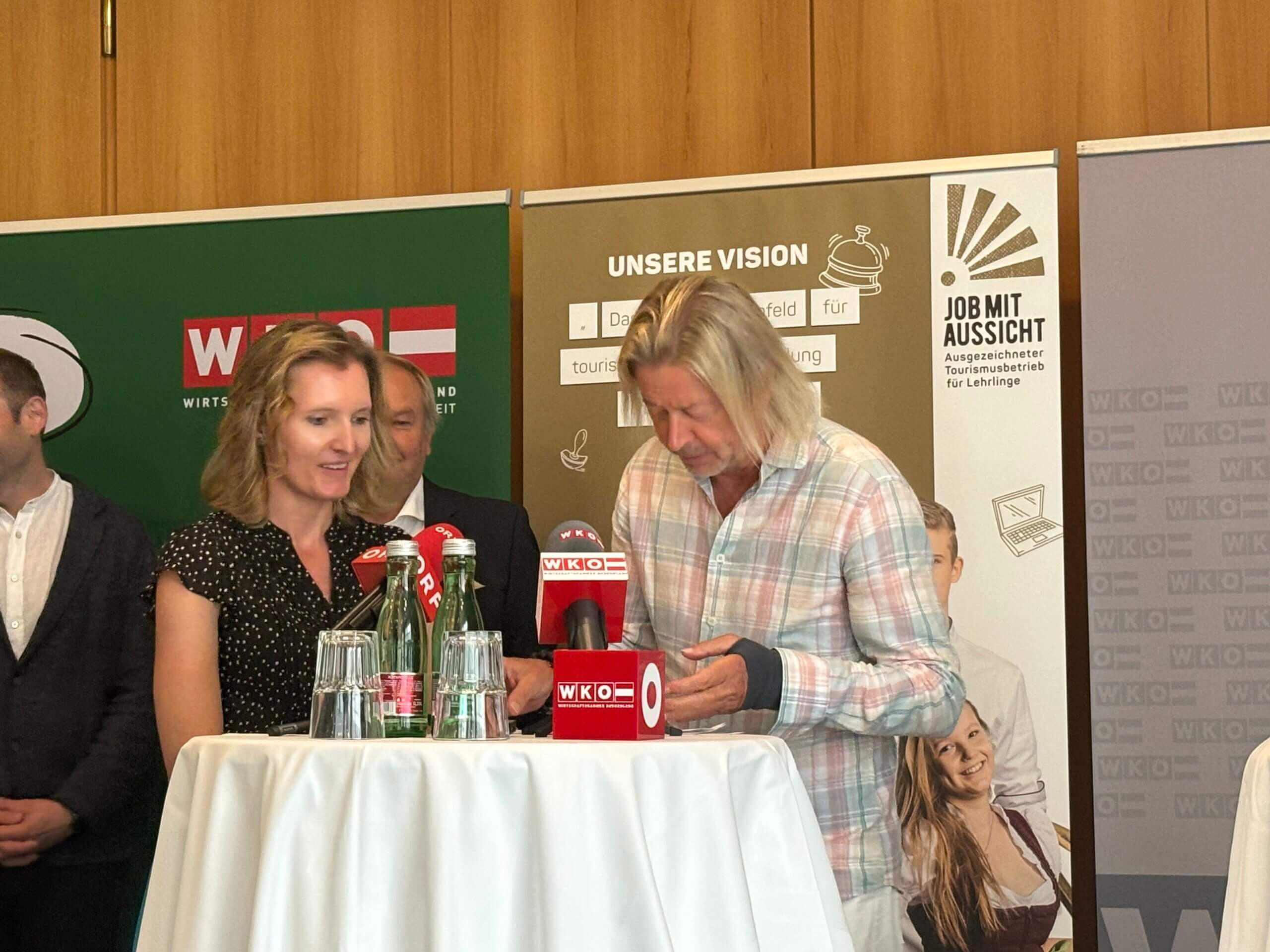 Karl Reiter und Marlies Rainer. Beide stehen hinter einem Tisch, auf dem mehrere Mikrofone platziert sind. Sie lächeln, während sie in die Mikrofone sprechen.