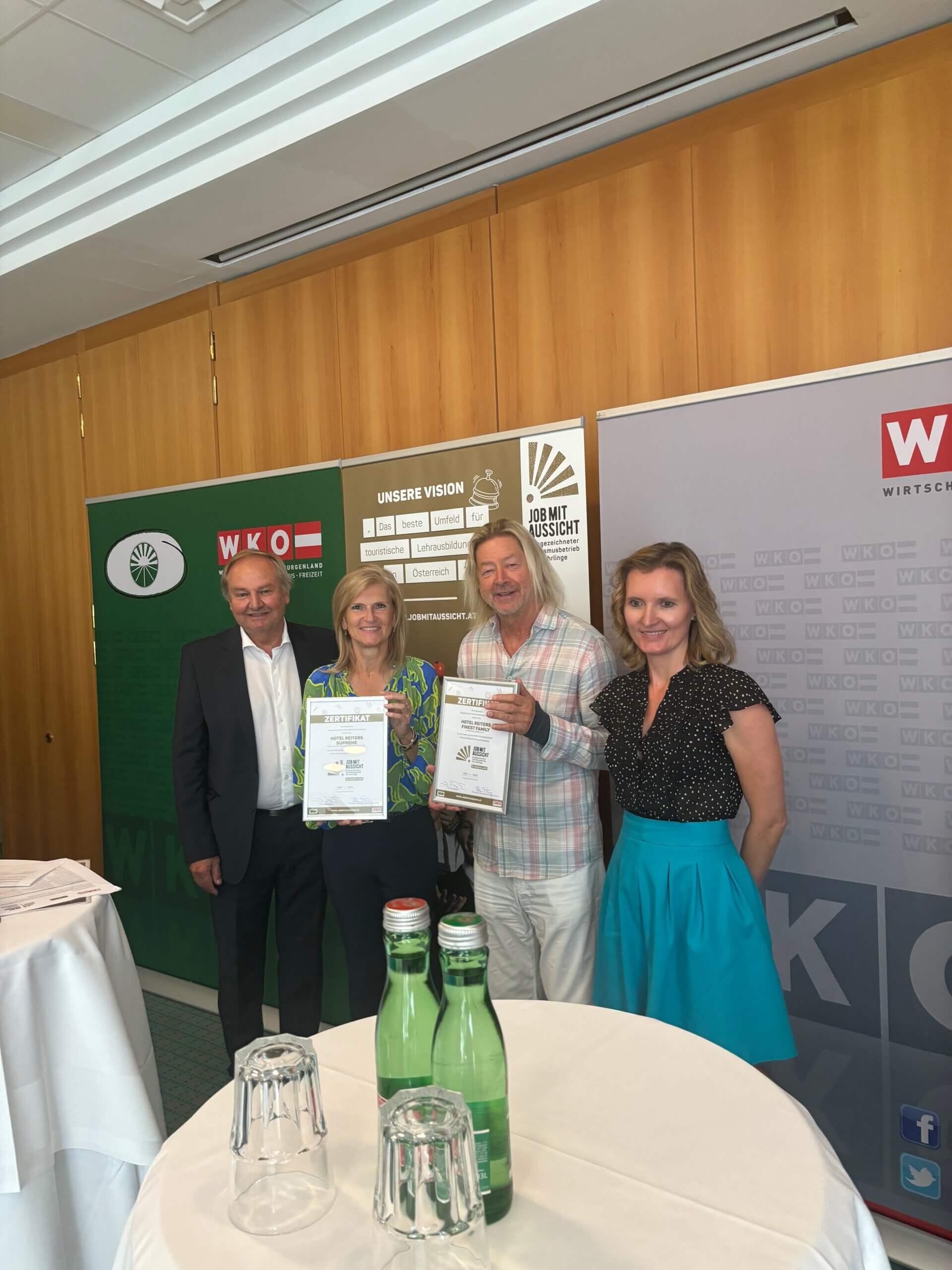Vier Personen stehen zusammen und halten Zertifikate in den Händen. Im Hintergrund sind Banner der Wirtschaftskammer Österreich (WKO) und eine weitere Organisation mit dem Slogan "JOB MIT AUSSICHT" zu sehen.