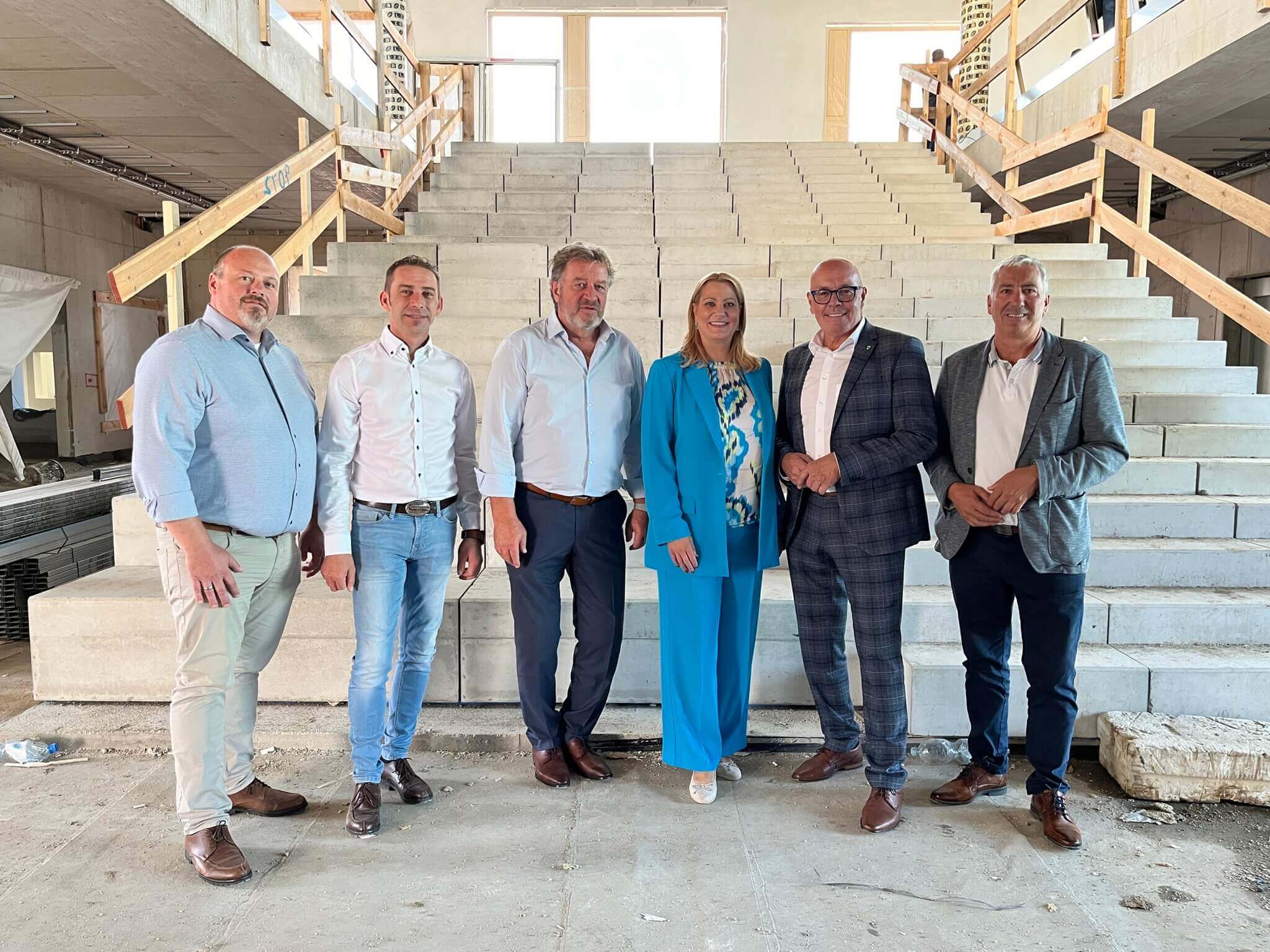 Im Vordergrund stehen sechs Personen nebeneinander vor einer breiten, unfertigen Betontreppe in einem Gebäude im Rohbau. Von links nach rechts: Amtsleiter Ing. Roland Poiger MBA, in einem hellblauen Hemd, beigen Hosen und braunen Schuhen; PEB-Geschäftsführer Thomas Rosner, in einem weißen Hemd, hellblauen Jeans und braunen Schuhen; Bildungsdirektor HR OSR Alfred Lehner BEd MA, in einem hellblauen Hemd, dunkelblauen Hosen und braunen Schuhen; LR Mag. (FH) Daniela Winkler, in einem leuchtend blauen Hosenanzug mit gemusterter Bluse; Bürgermeister Georg Rosner, in einem grauen karierten Anzug mit weißem Hemd und Brille; und Vizebürgermeister Michael Leitgeb, in einem grauen Blazer, weißem Polohemd, dunkelblauen Hosen und braunen Schuhen. Im Hintergrund ist ein großer, unfertiger Innenraum mit rohen Betonwänden und hölzernen Geländern an der Treppe zu sehen. Foto: Stadtgemeinde Oberwart