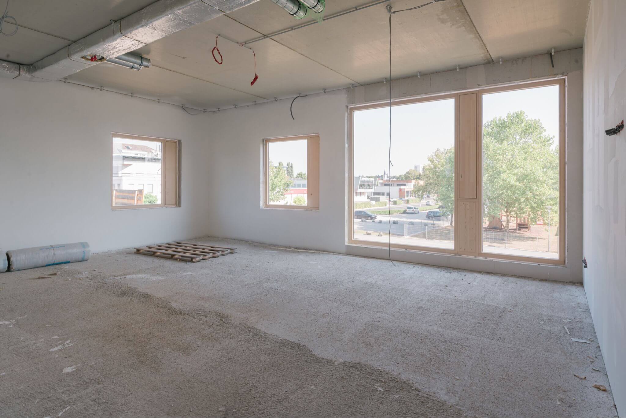 Ein unfertiger Raum in einem Rohbau. Der Boden ist mit grobem Beton bedeckt, und an der Decke verlaufen sichtbare Lüftungsrohre sowie lose hängende Kabel. Auf dem Boden liegen eine Holzpalette und eine aufgerollte Schutzfolie. An der Rückwand befinden sich zwei rechteckige Fenster, durch die Tageslicht einfällt. Auf der rechten Seite des Raums ist ein großes bodentiefes Fenster mit Blick auf eine Straße und einige Bäume zu sehen. Im Außenbereich sind geparkte Autos und Gebäude in der Ferne erkennbar. (c) Franz&Sue/Louai Abdul Fattah