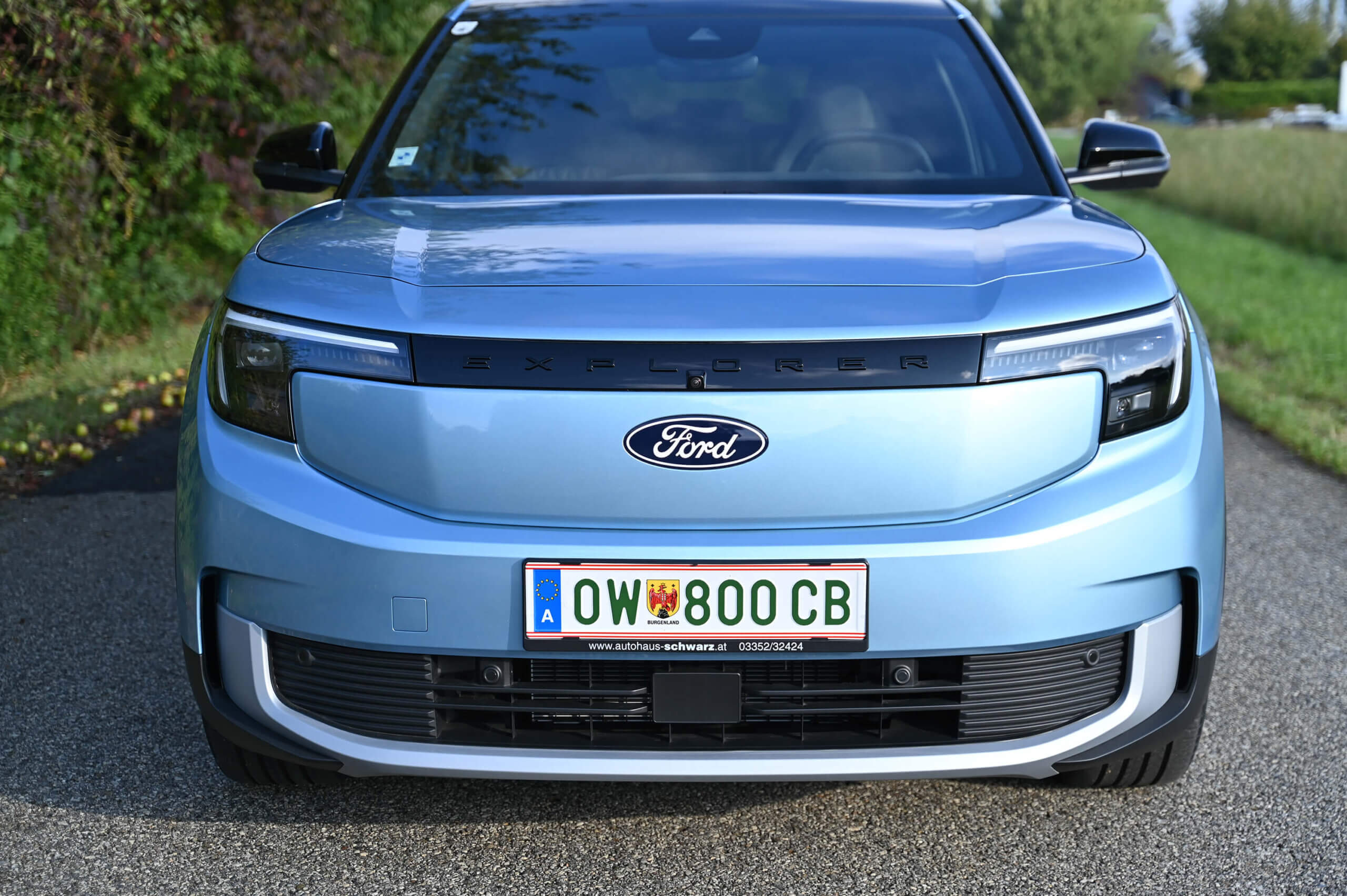 Die Front eines hellblauen Ford Explorer ist zu sehen. Auffällig ist der schwarze Schriftzug „EXPLORER“ über dem Ford-Logo in der Mitte des Kühlergrills. Die LED-Scheinwerfer haben ein modernes, eckiges Design. Unten ist ein österreichisches Nummernschild mit der Kennung „OW 800 CB“ zu erkennen, wobei „OW“ für Oberösterreich steht. Das Fahrzeug steht auf einer asphaltierten Straße, während im Hintergrund links Grünflächen und Bäume zu sehen sind. Rechts erstreckt sich eine Wiese neben der Straße. Foto: Eva Maria Kamper