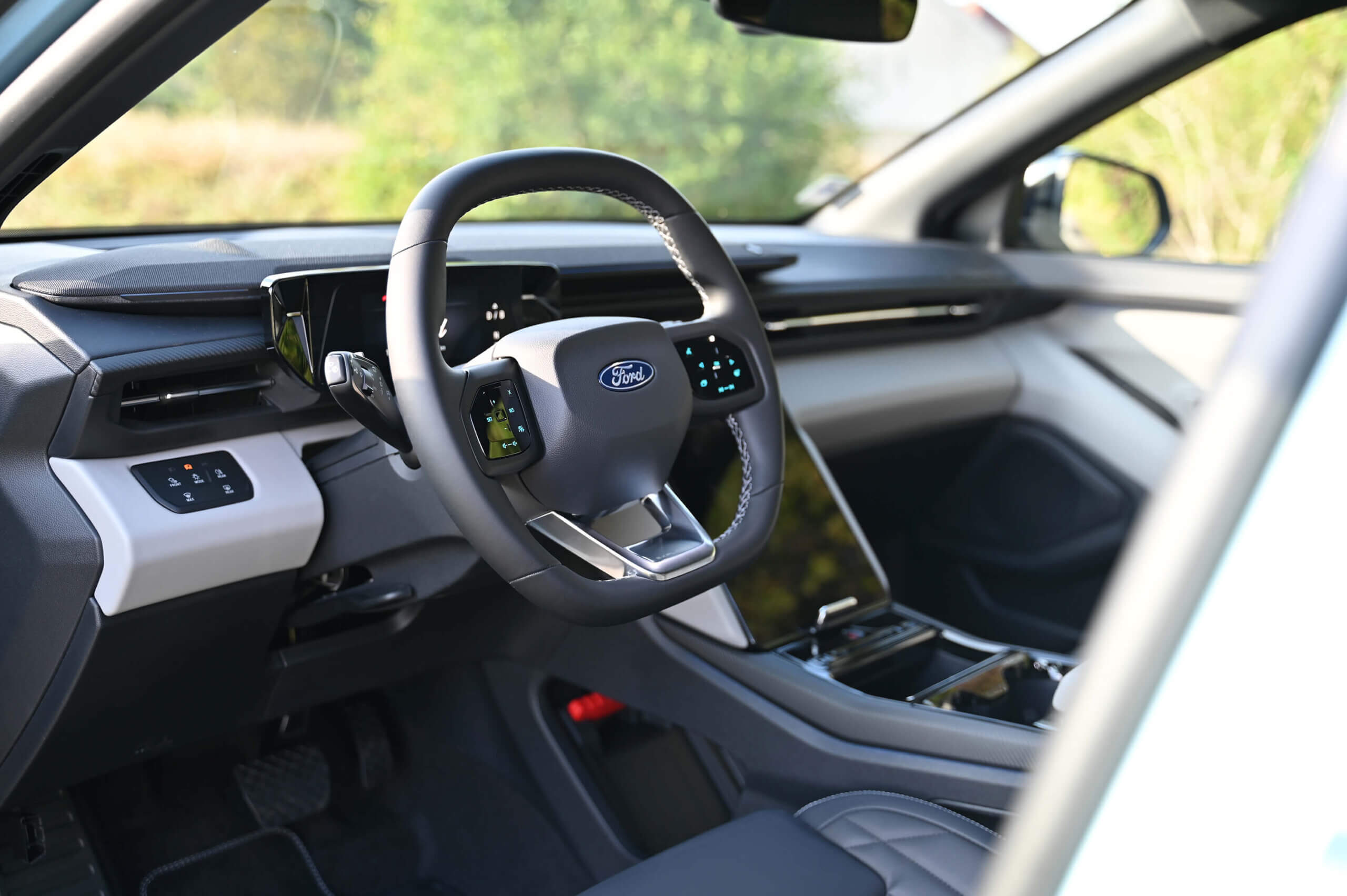 Das Innere des Ford Explorer zeigt ein modernes, minimalistisches Design. Im Fokus steht das Lenkrad mit Ford-Logo in der Mitte, das auf beiden Seiten Touch-Bedienelemente integriert hat. Hinter dem Lenkrad befindet sich ein digitales Display als Instrumententafel. Die Mittelkonsole ist mit einem großen Touchscreen ausgestattet, der in den schwarzen, glänzenden Armaturen integriert ist. Das Cockpit und der Innenraum sind in Grau- und Schwarztönen gehalten, mit kontrastierenden Nähten auf den Sitzen. Die Umgebung außerhalb des Autos ist durch die Seitenfenster sichtbar. Foto: Eva Maria Kamper